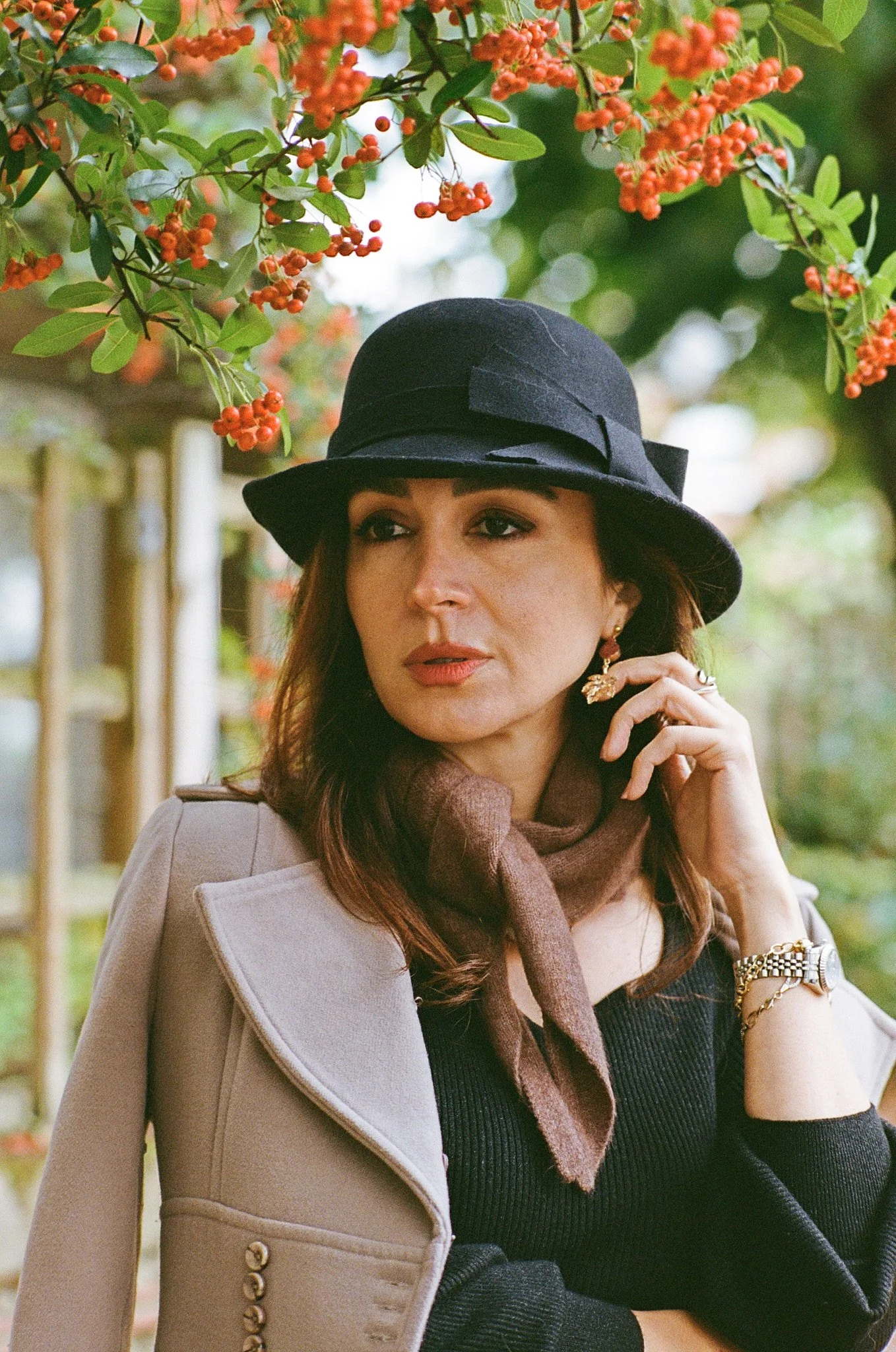 A woman in a black hat and beige coat is standing outdoors surrounded by greenery and orange-red berries, with her hand near her face.