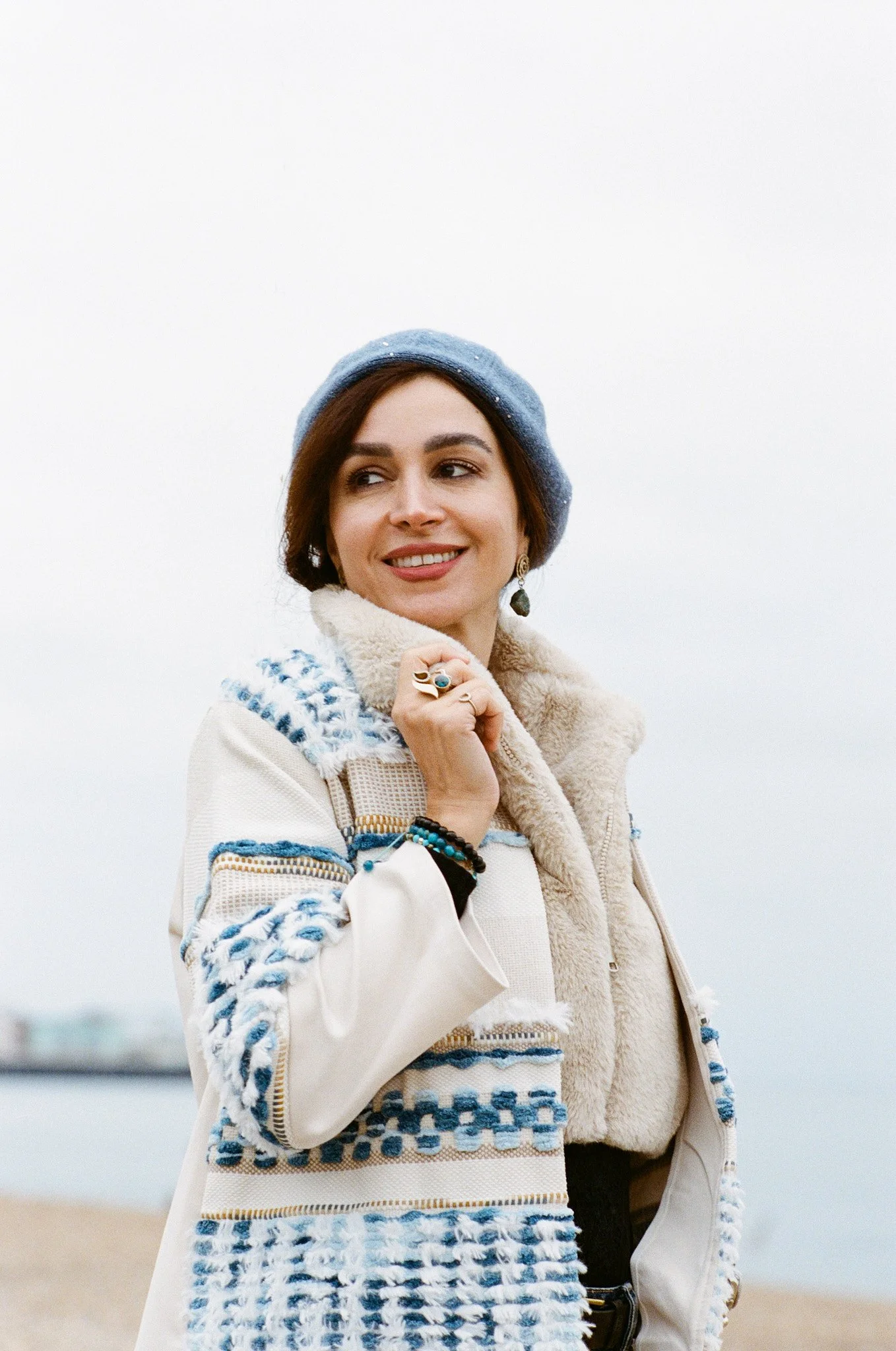 A woman outdoors on a cloudy day, wearing a blue beret, a beige and blue woven jacket with a shearling collar, and jewelry, smiling and looking away.