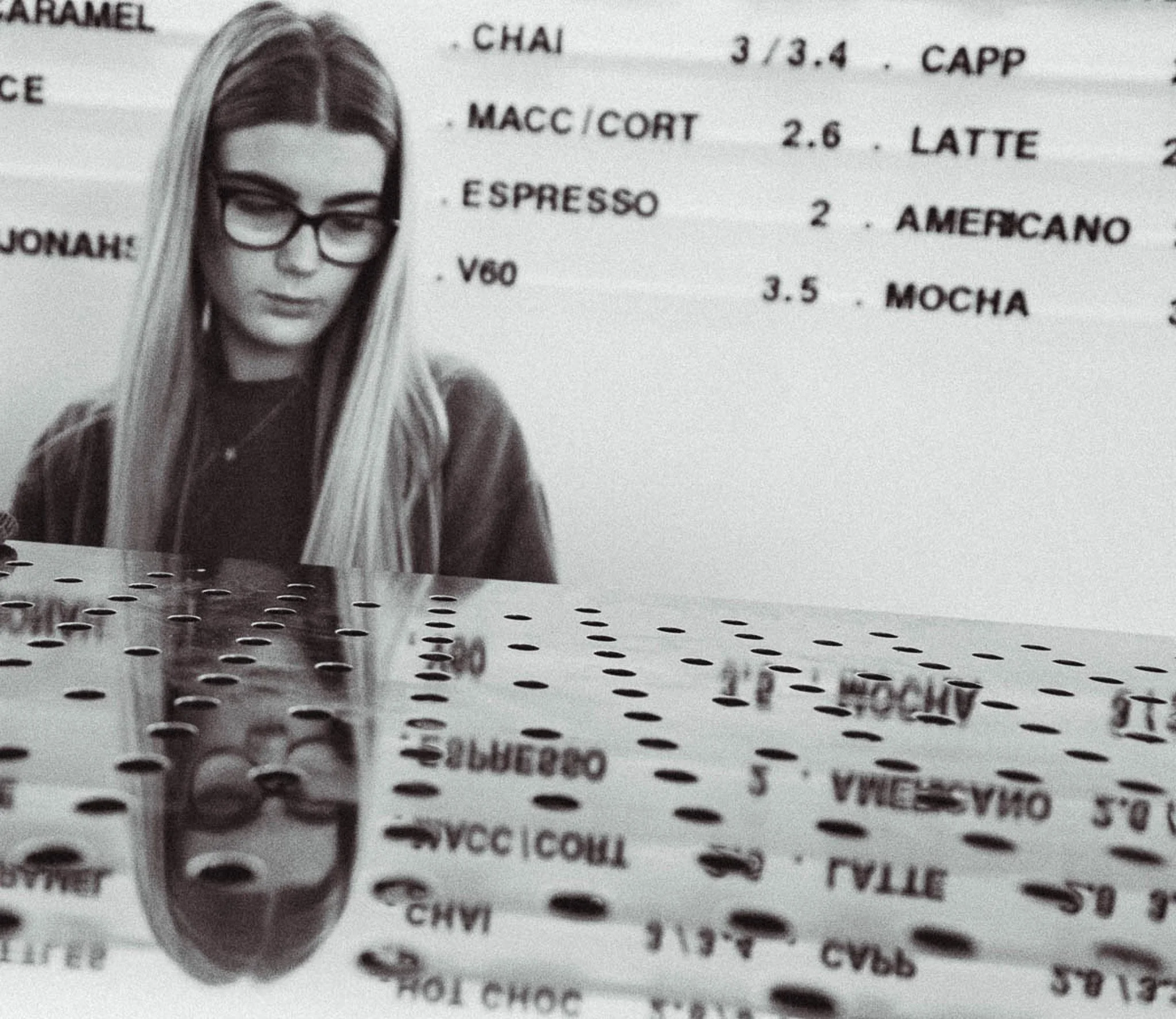Black and white photo of a girl with long straight hair wearing glasses and a dark top, standing next to a menu board with coffee options and prices.