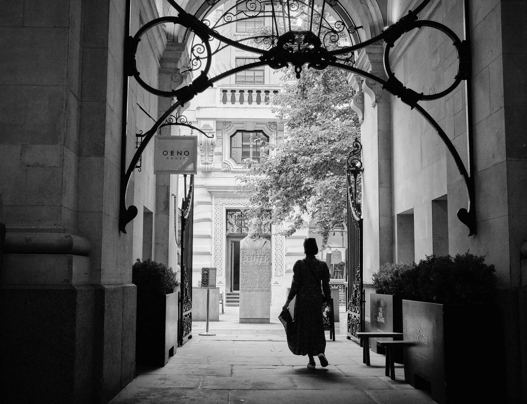 A person walks through an ornate iron gate into a courtyard with classical architecture, a statue, and a sign that reads 'Pauli Juus Reuter' in black and white.