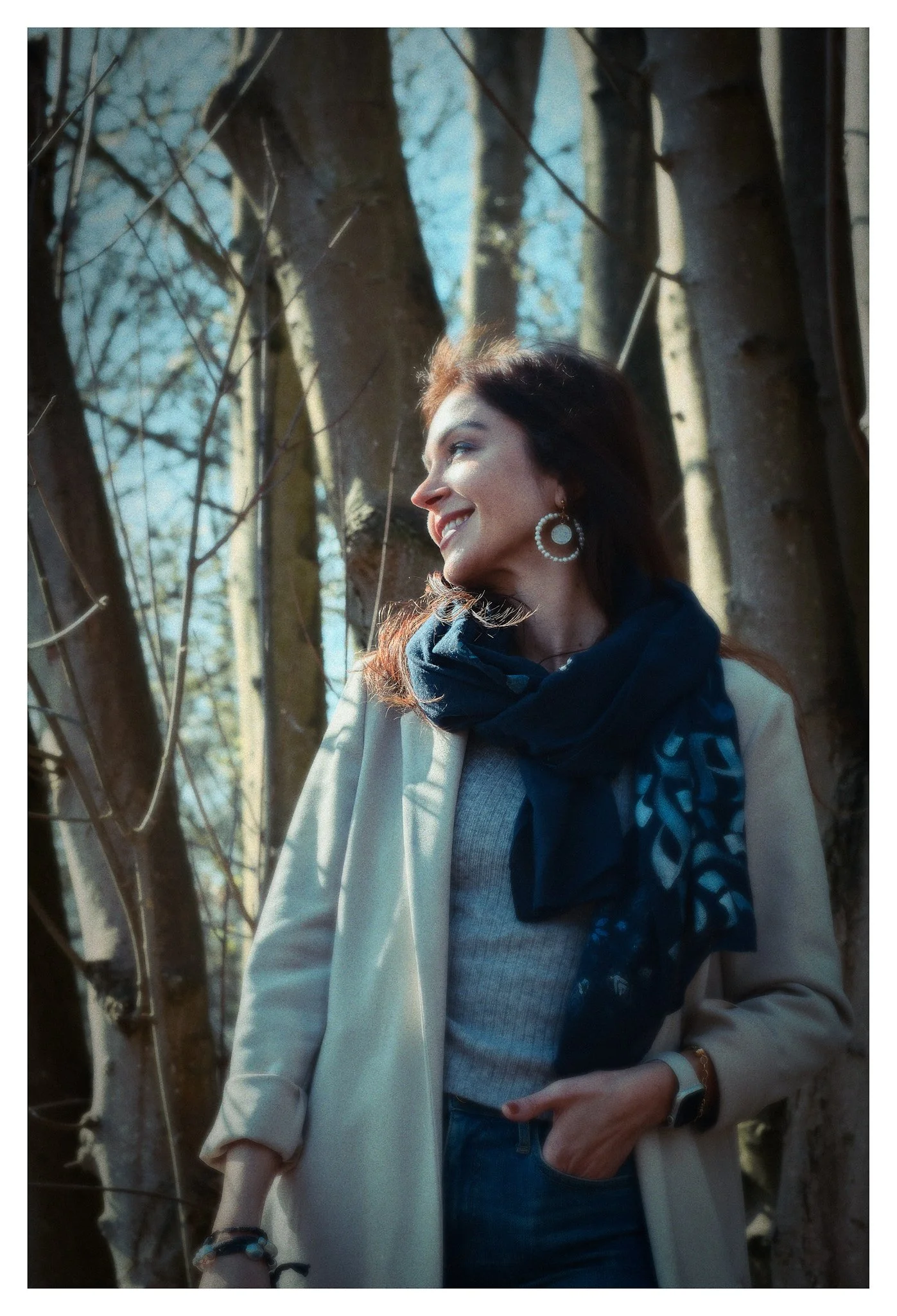 Woman smiling outdoors among tree trunks, wearing earrings, a blue scarf, gray top, beige coat, and accessories.