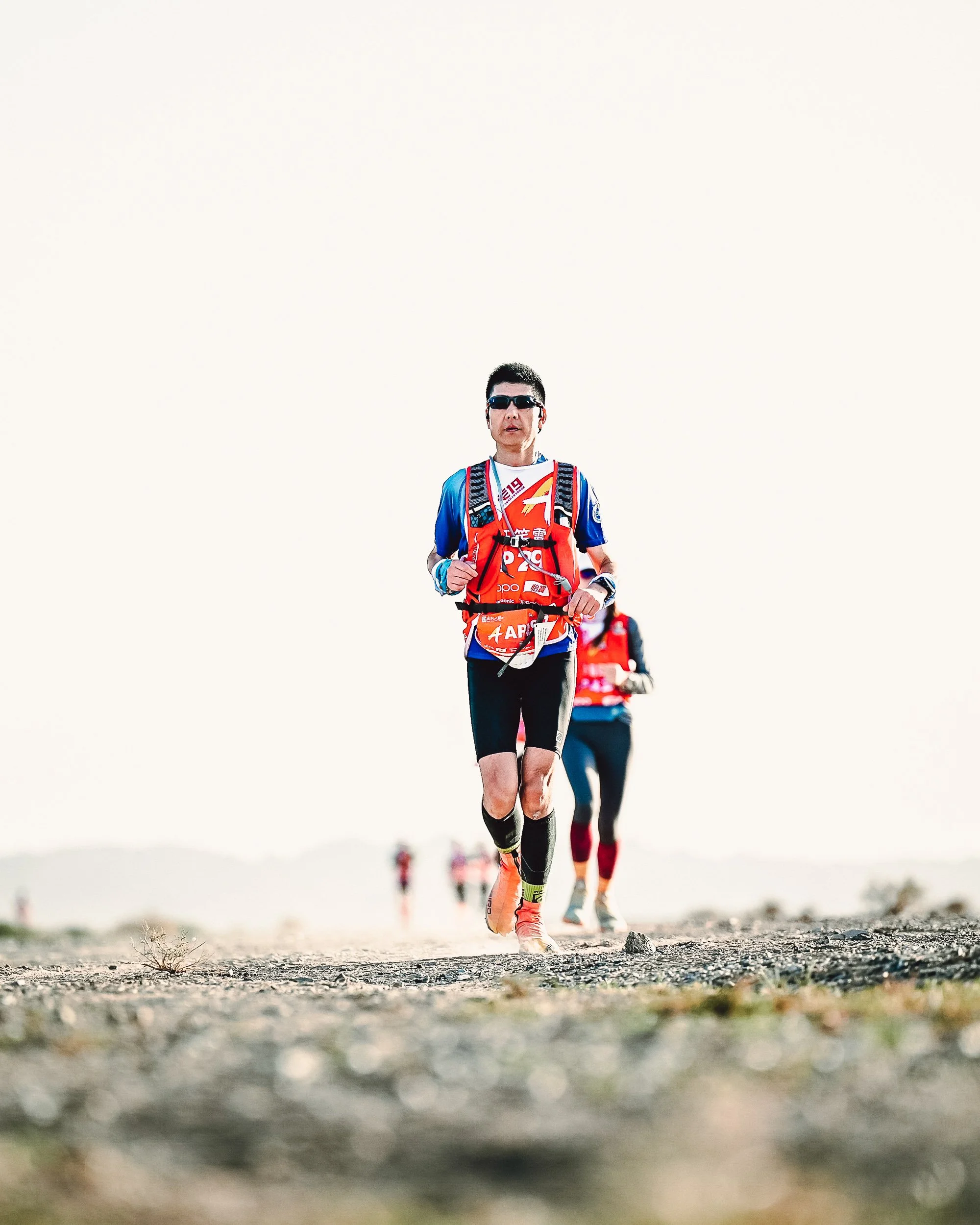 A male trail runner wearing sunglasses, a race bib, and a red hydration vest runs on a rocky terrain with a group of runners behind him under a pale sky.