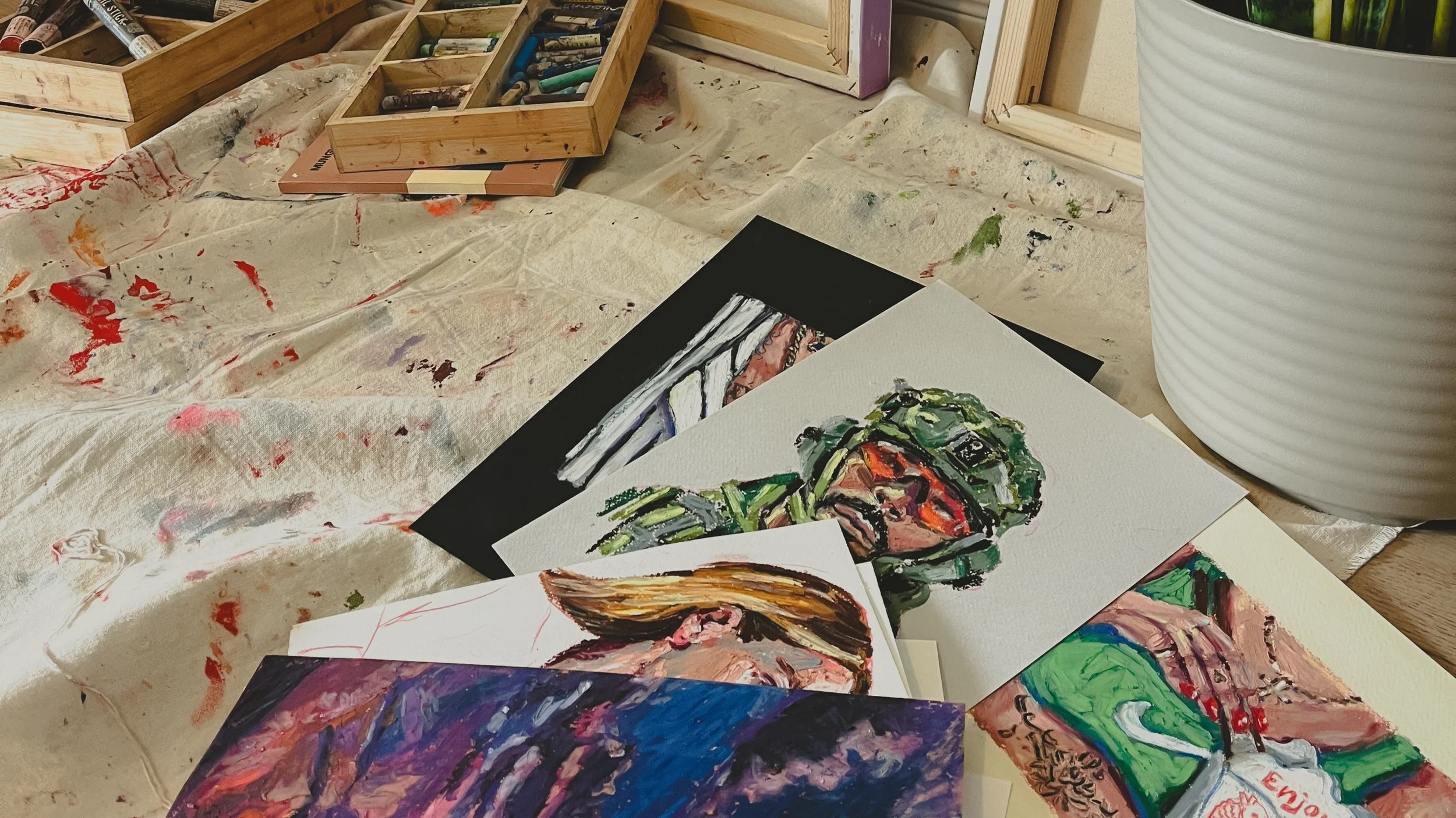 A workspace with various painted portrait cards, paint splatters on a cloth, and painting supplies in boxes. One plant pot is partially visible.