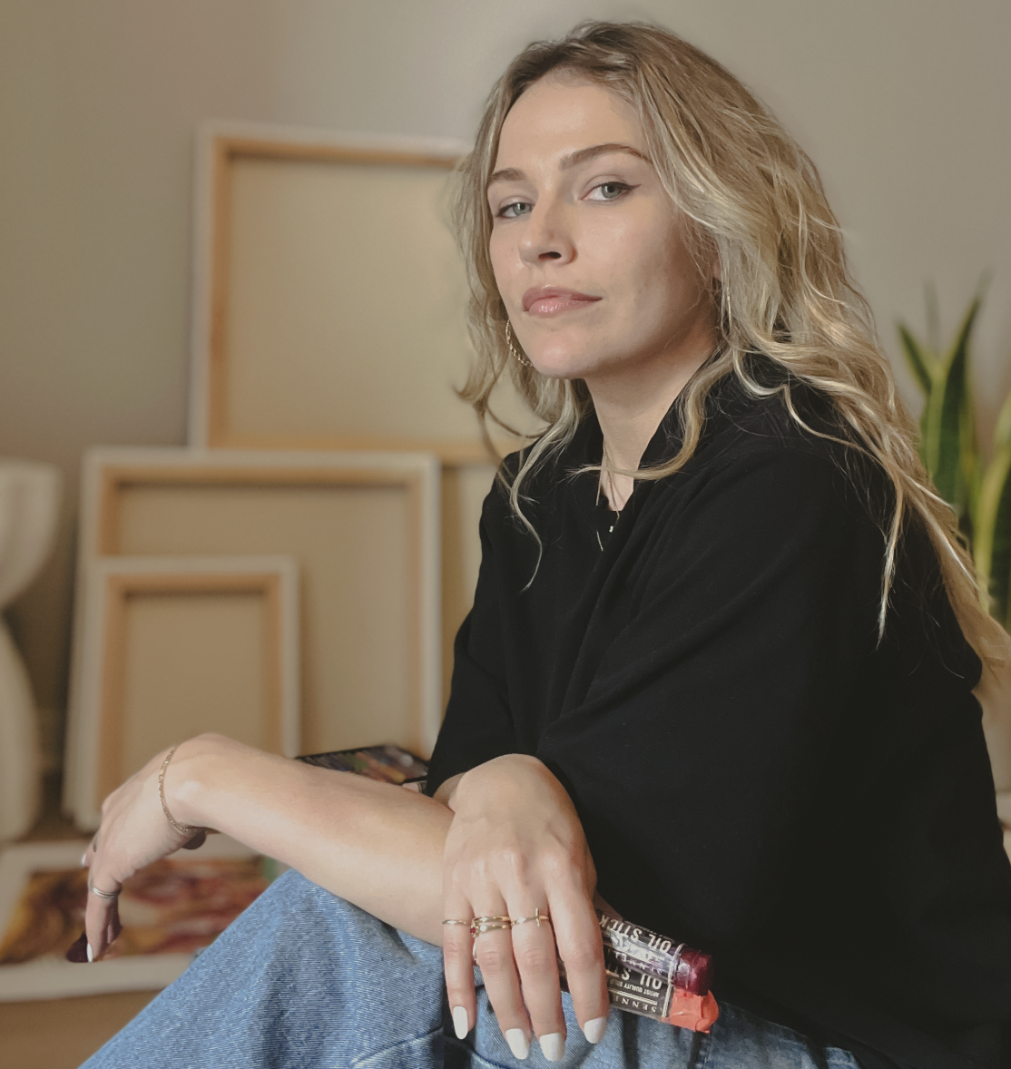 A woman with wavy blonde hair and light skin sitting indoors in her studio, holding oil pastels, with empty picture frames and a plant in the background.