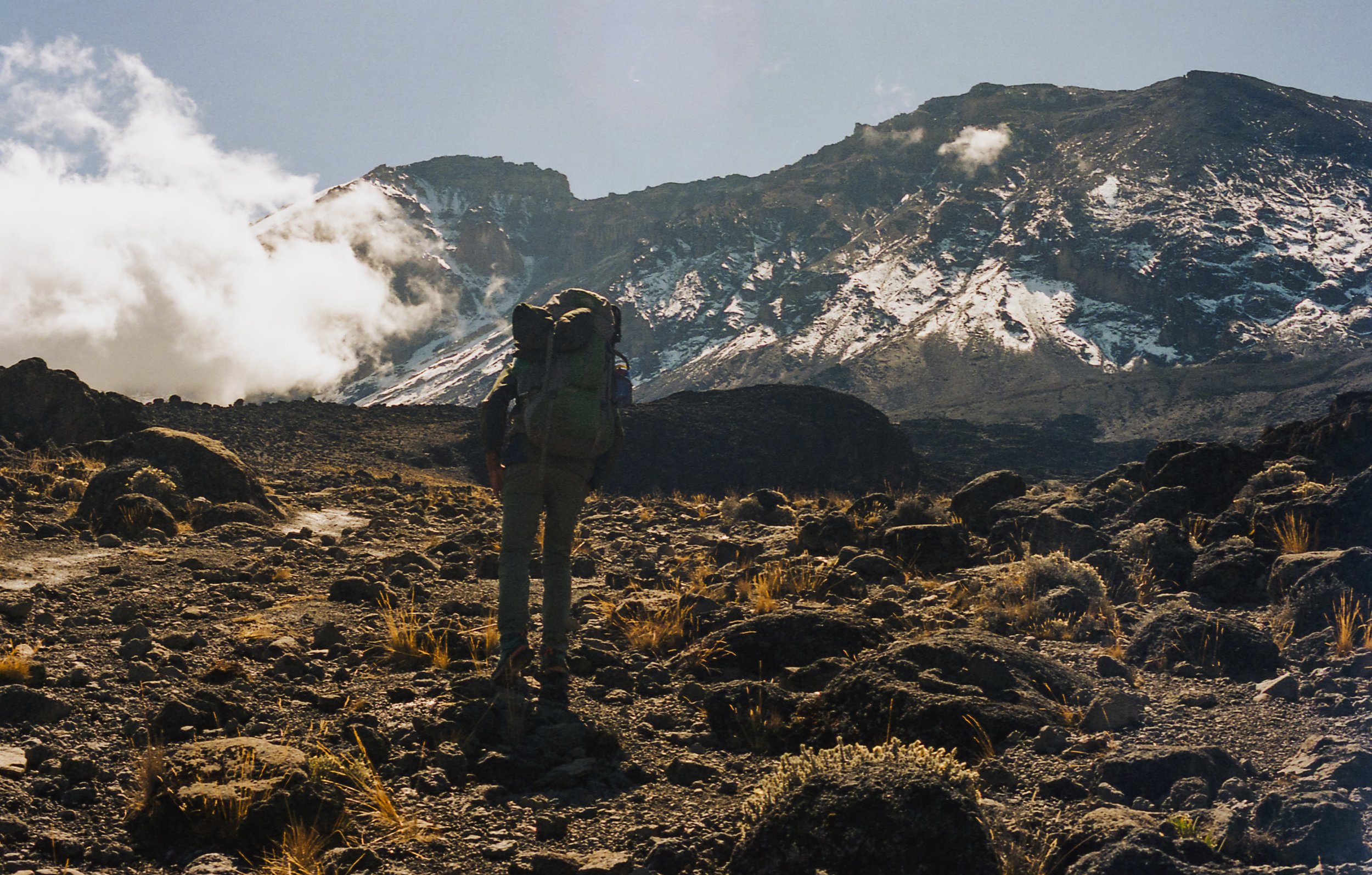 A lone hiker with a large backpack trekking through a rocky volcanic landscape with snow-capped mountains in the background and clouds surrounding the peaks.