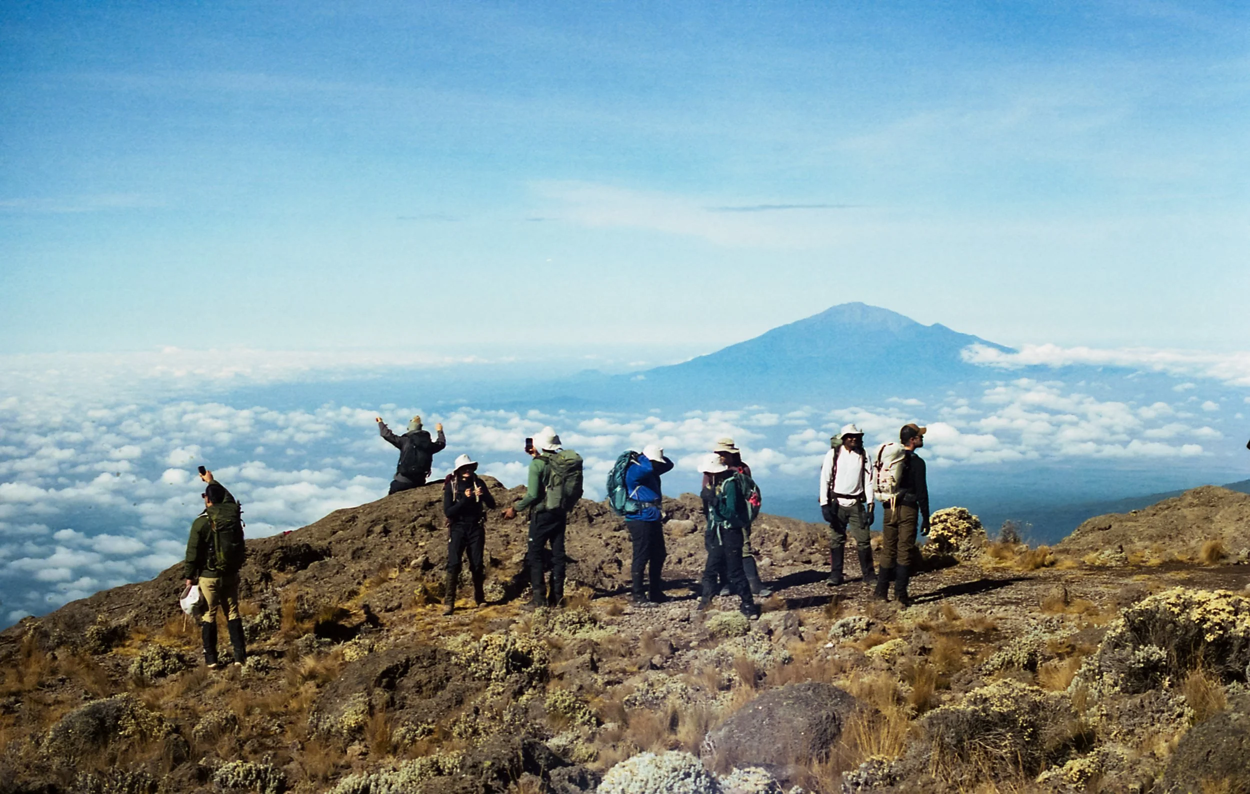 Group of hikers on a mountain summit with clouds and a distant volcano in the background.