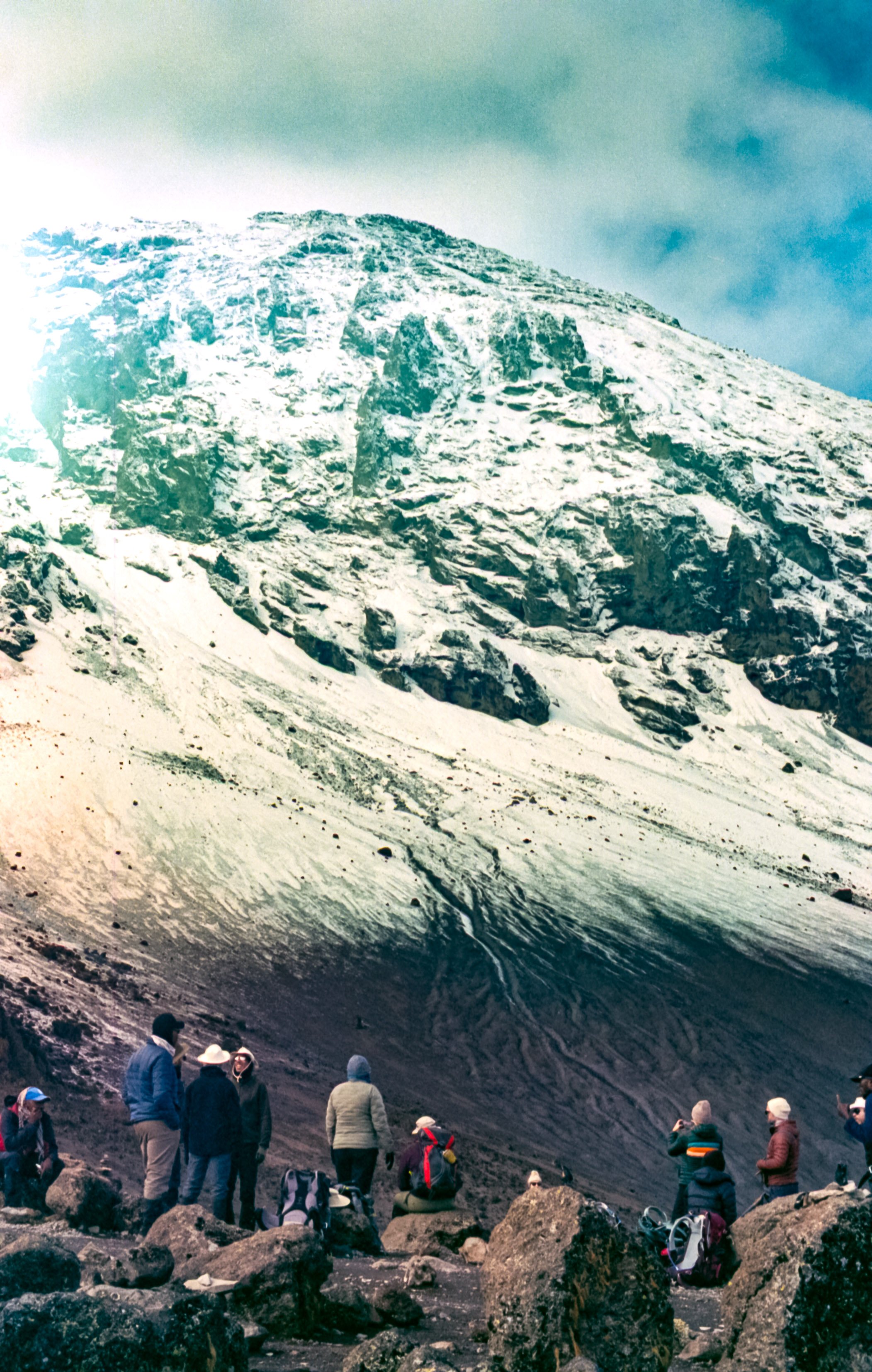 Group of hikers resting at the base of a snow-covered mountain, with rocky terrain and cloudy sky.