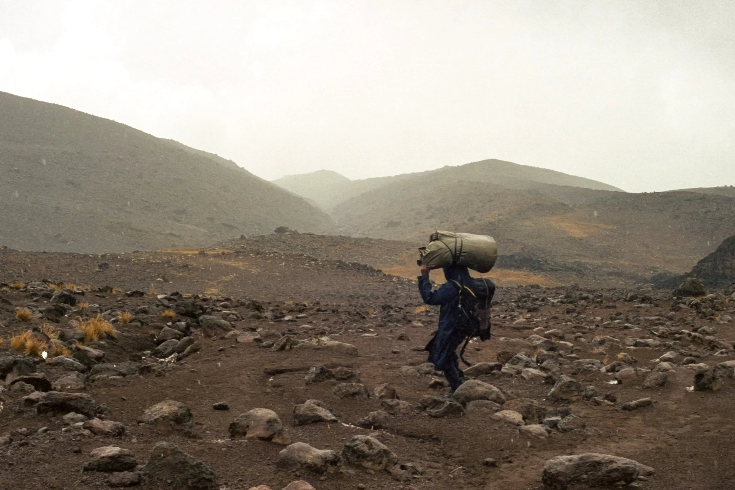 Hiker with a large backpack walking across a rocky, barren landscape with mountains in the background on a cloudy day.