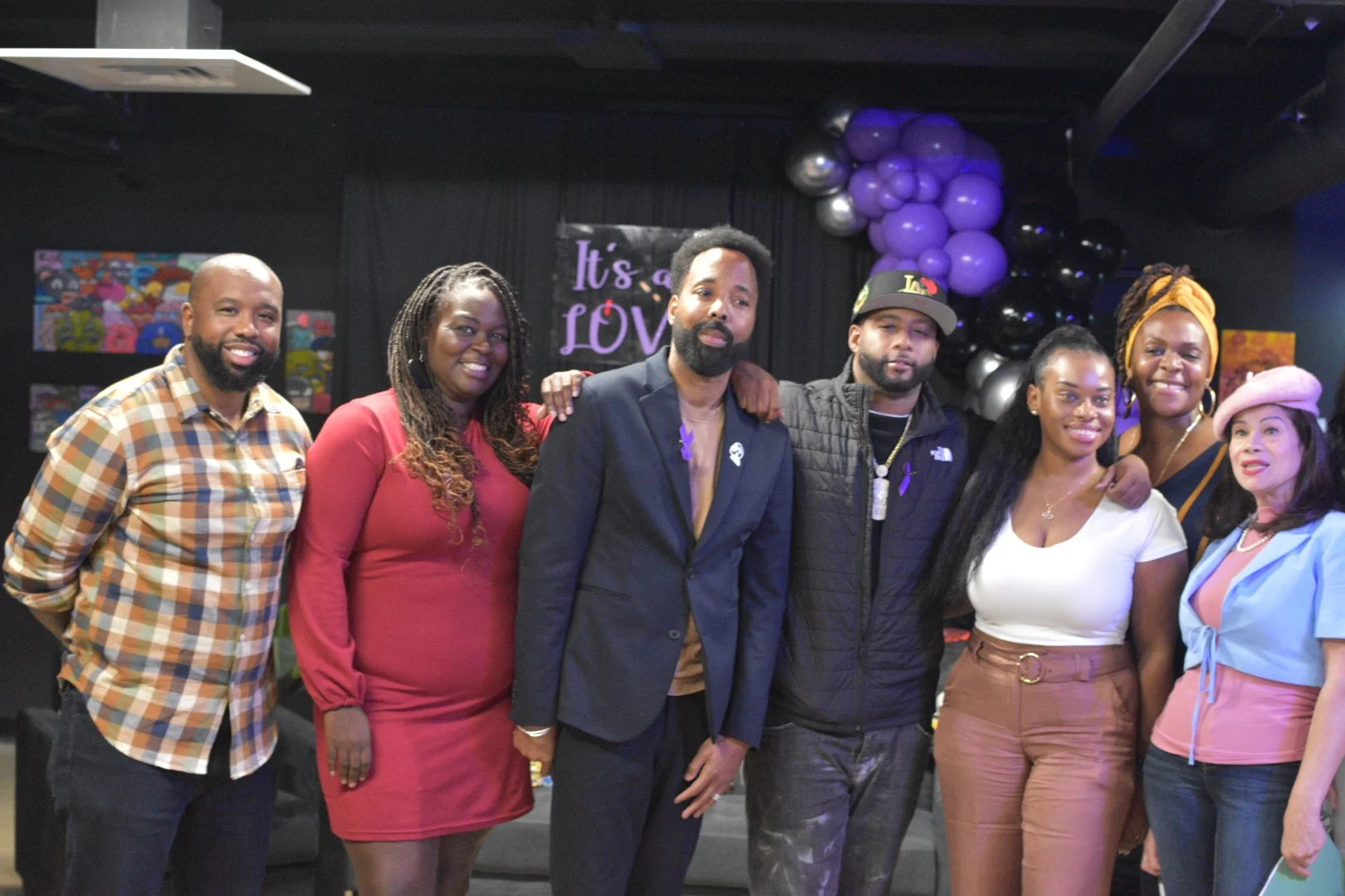 A group of seven people posing close together indoors, with background decorations including purple balloons and a "It's a Love" sign. The individuals are wearing a mix of casual and semi-formal attire.