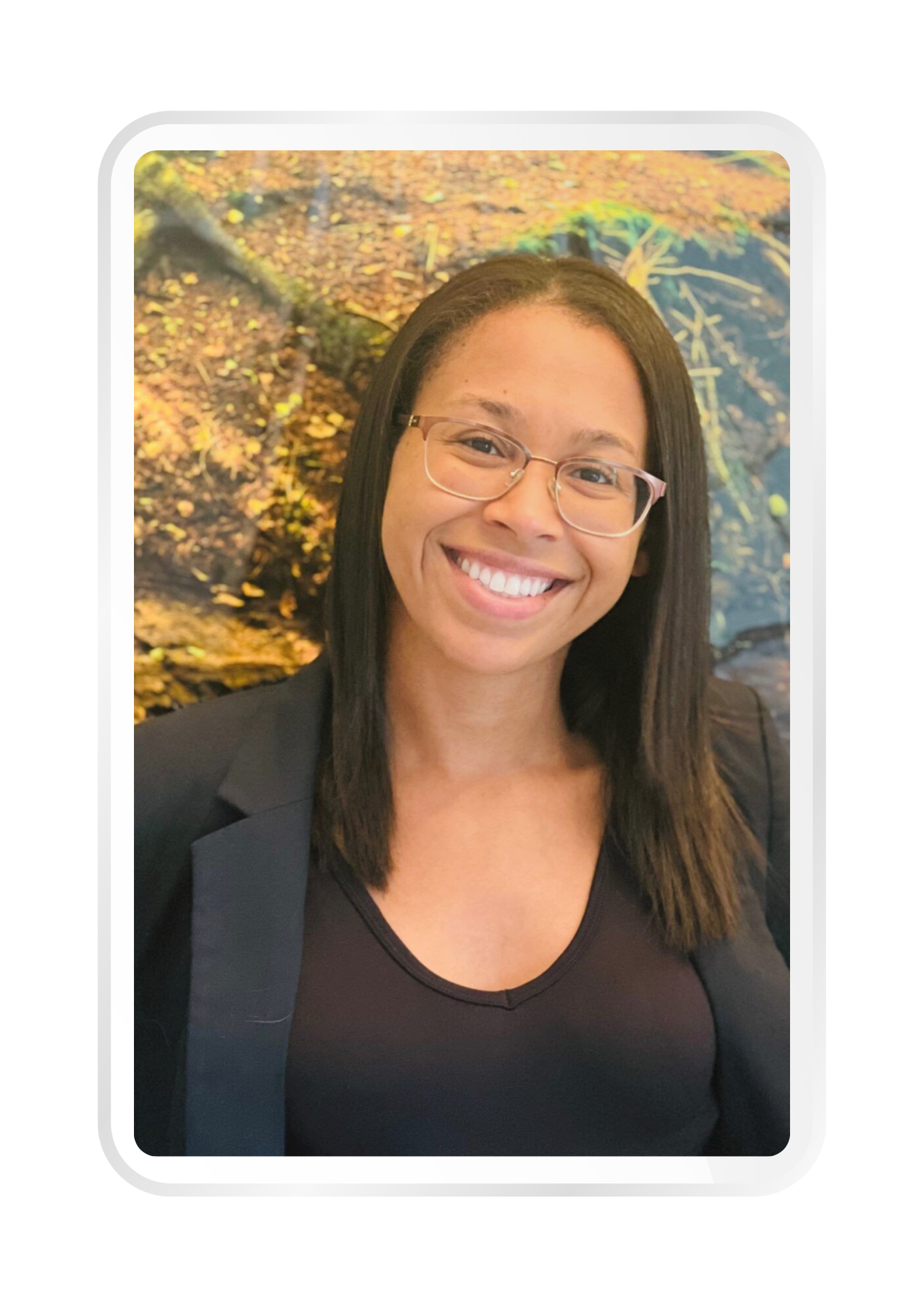 A smiling person with glasses wearing a black blazer and shirt, standing in front of a nature-themed background.