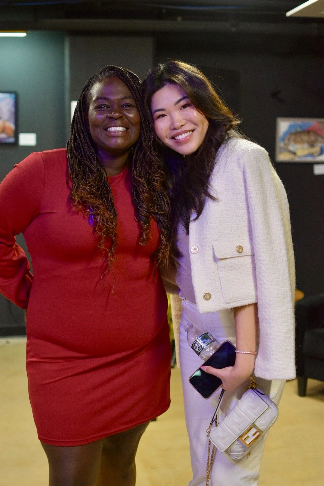 Two smiling women posing in a casual indoor setting, one wearing a red dress and the other in a white jacket and pants, holding a phone and a small white handbag.