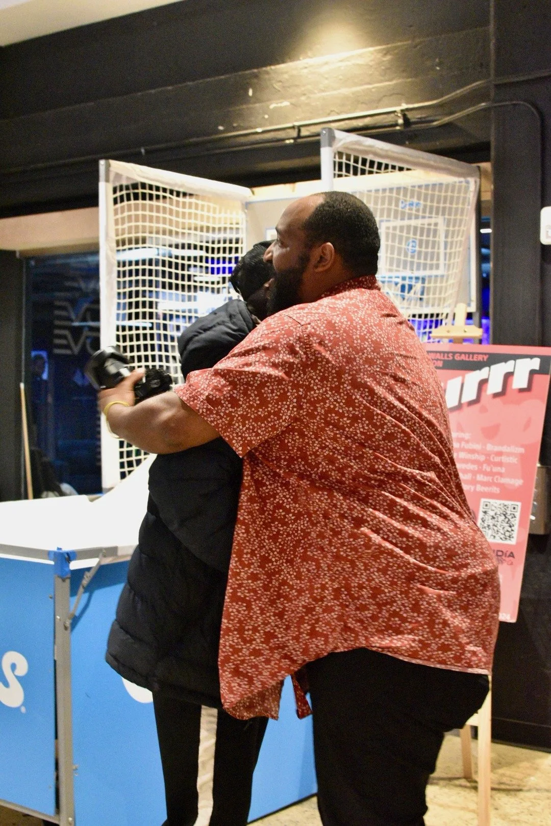 Two people hugging in an indoor space with netted structures and a red patterned sign in the background.