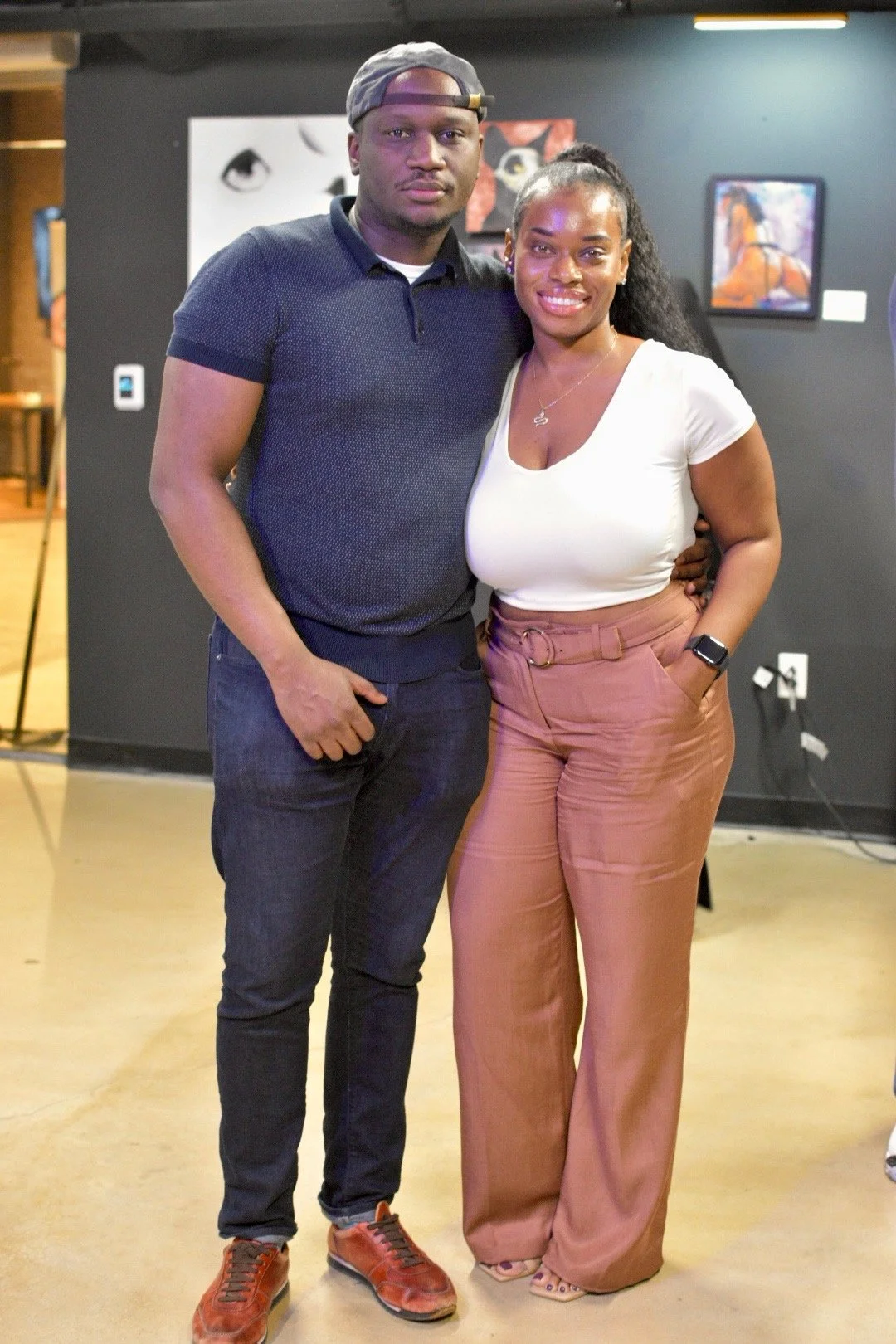 A man and woman smiling and posing together indoors, with artwork displayed on the wall behind them. The man is wearing a cap and dark outfit, while the woman is wearing a white top and pink pants.