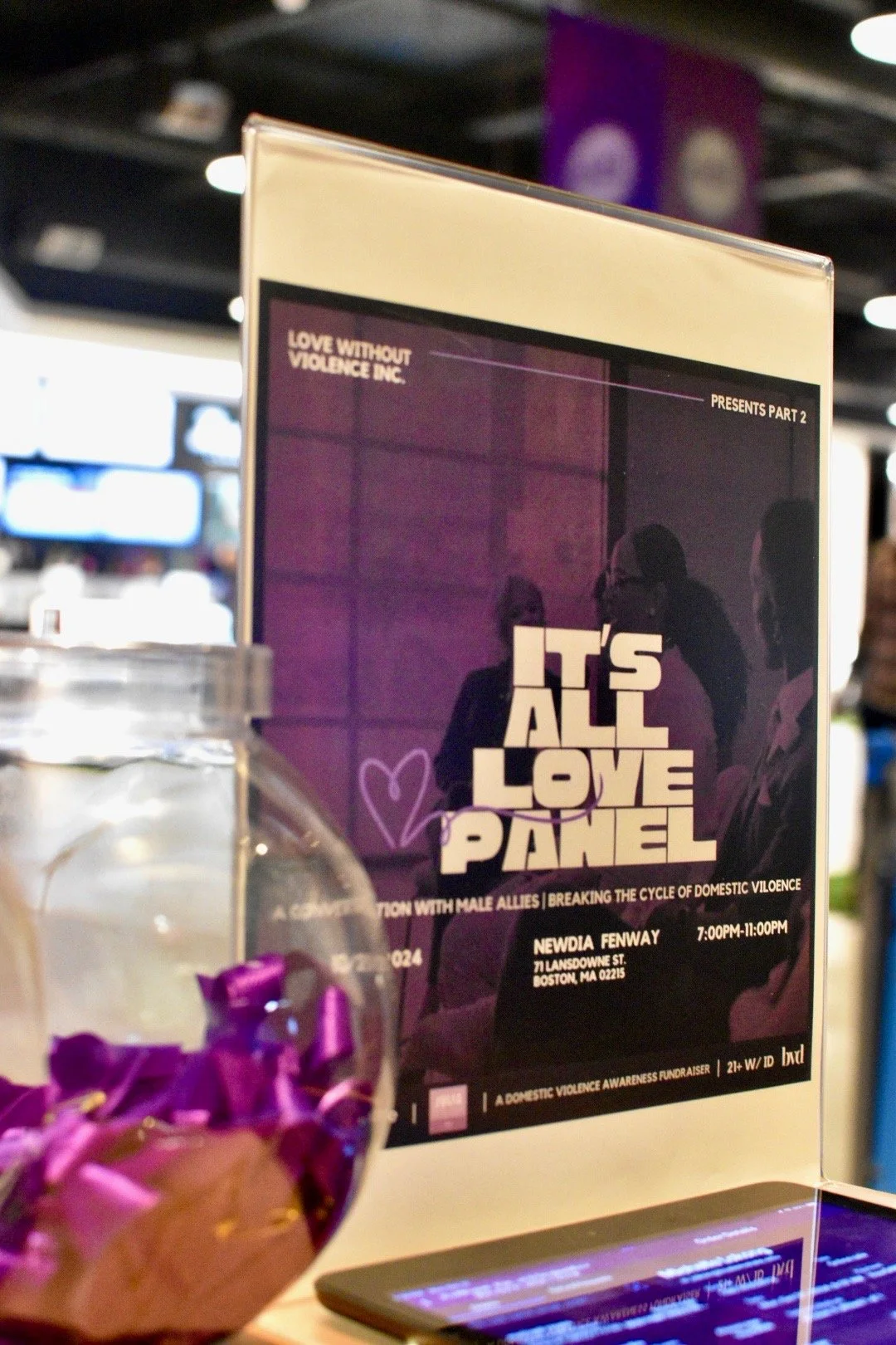 Promotional display for 'It's All Love' panel, featuring event details on breaking the cycle of domestic violence, with a jar of purple ribbons beside it.