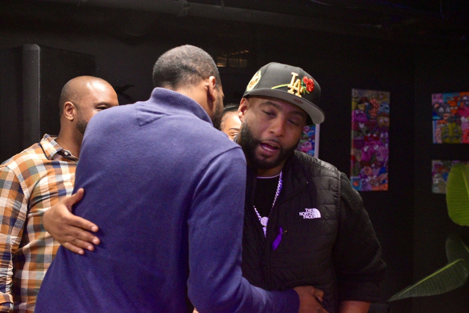 Group of people in a casual setting, two men embracing, one wearing a cap, quilted jacket, and chain, with colorful artwork in the background.