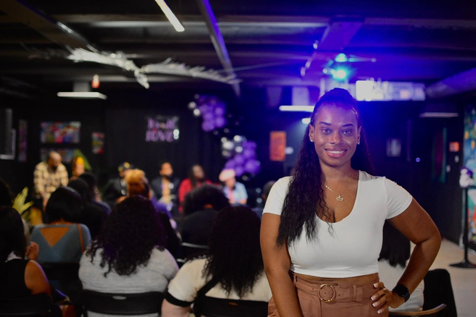 Woman smiling in a crowded room with people seated in the background, out of focus, suggesting a conference or event.