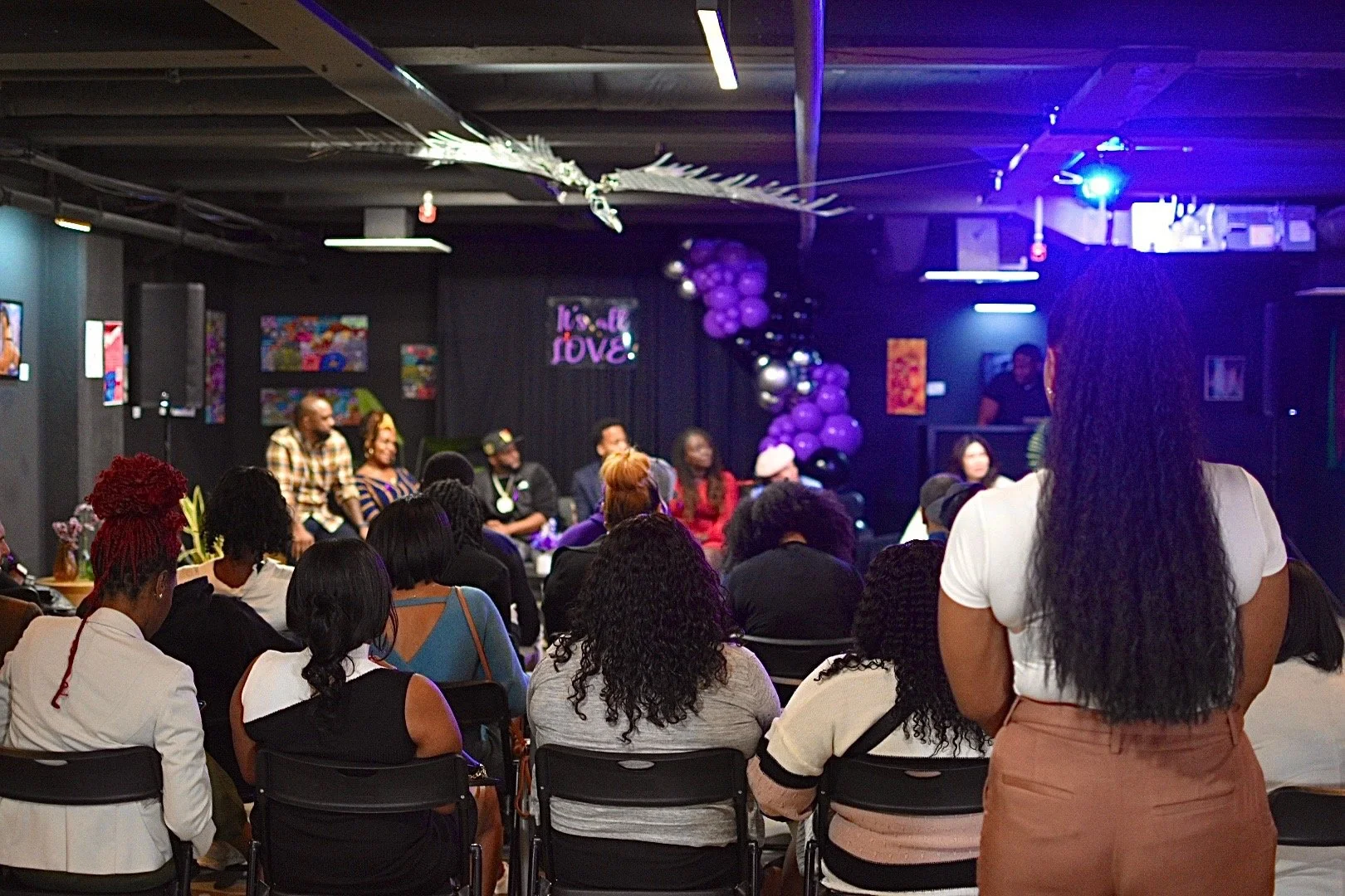 Audience attending an indoor panel discussion with a group of speakers on stage, decorated with purple and black balloons, in a room with colorful artwork on the walls.