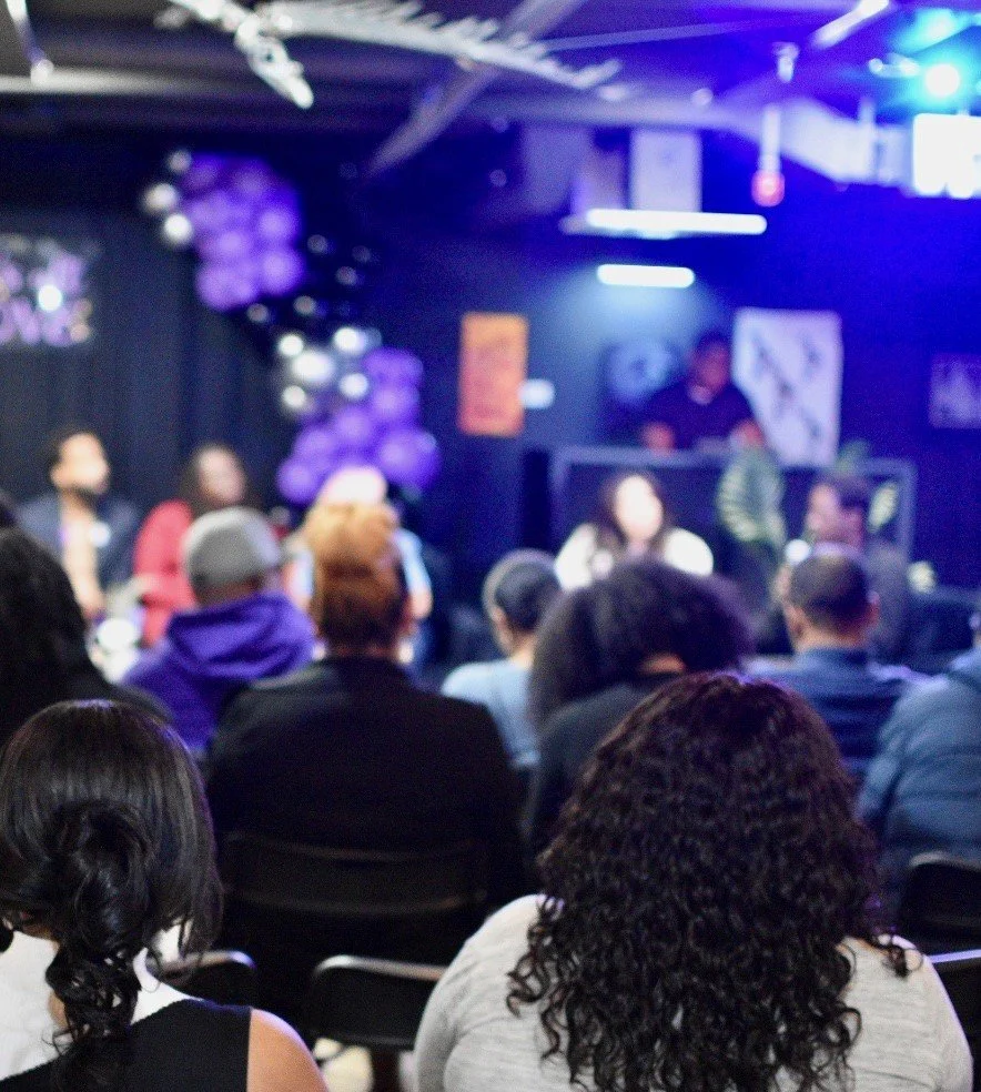 Audience watching a panel discussion in a dimly lit room with purple and black balloons.