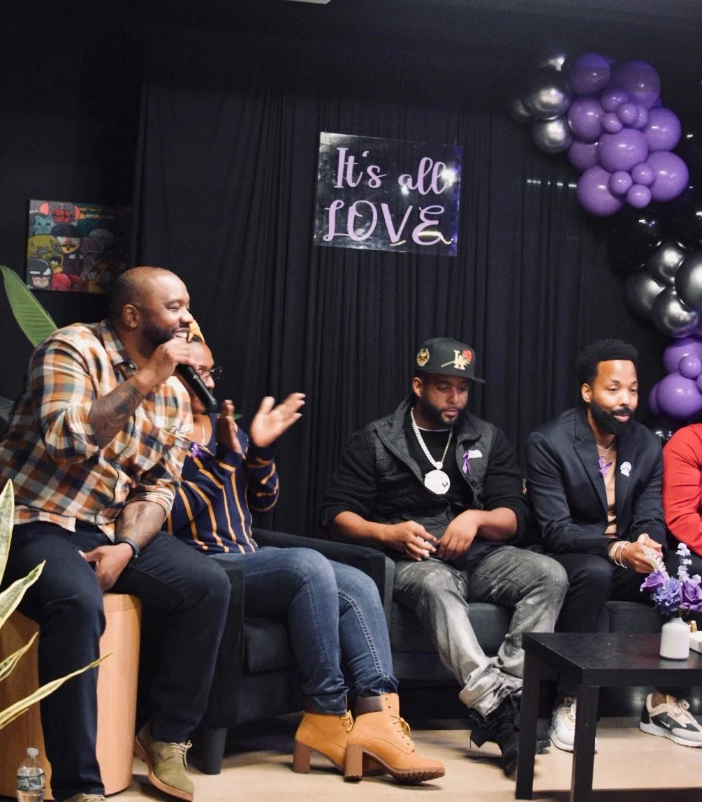 A group of people sitting on a couch engaged in a discussion, with a "It's all LOVE" sign behind them. There are purple and black balloons decorating the space. One person is holding a microphone, indicating a conversation or a panel event.