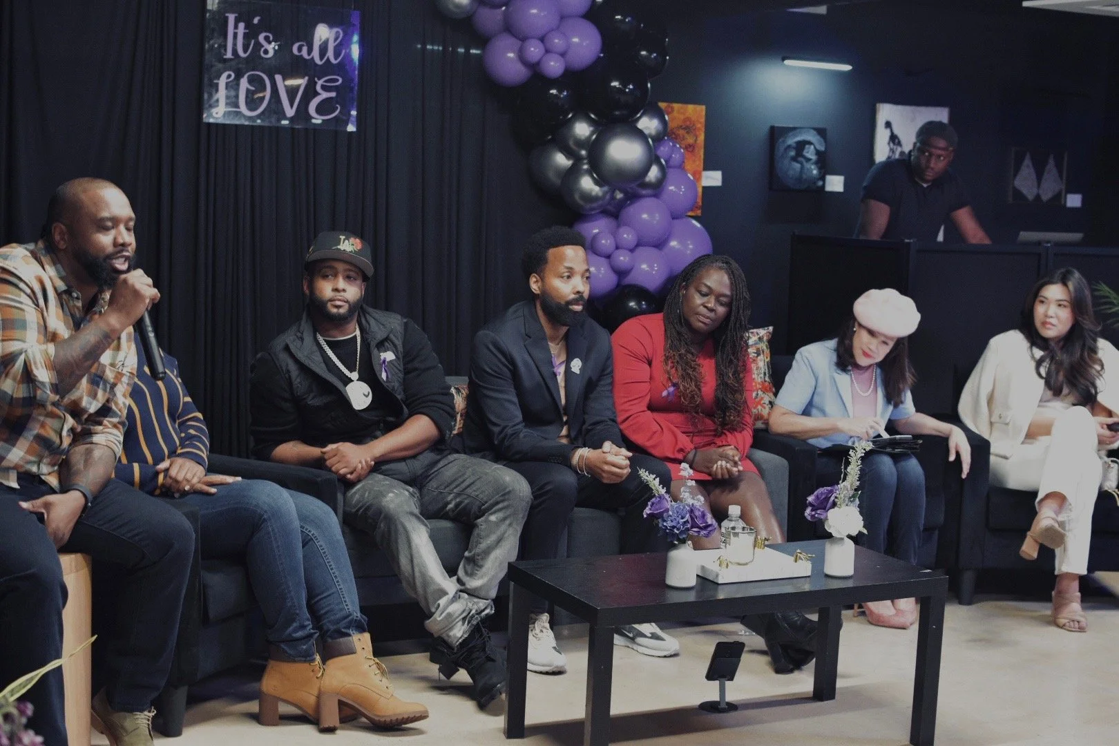 A group of people sitting together in a seminar or discussion setting, with a "It's All Love" sign in the background and purple and black balloons. The individuals, some holding microphones, are participating in an event. A table with decorative item