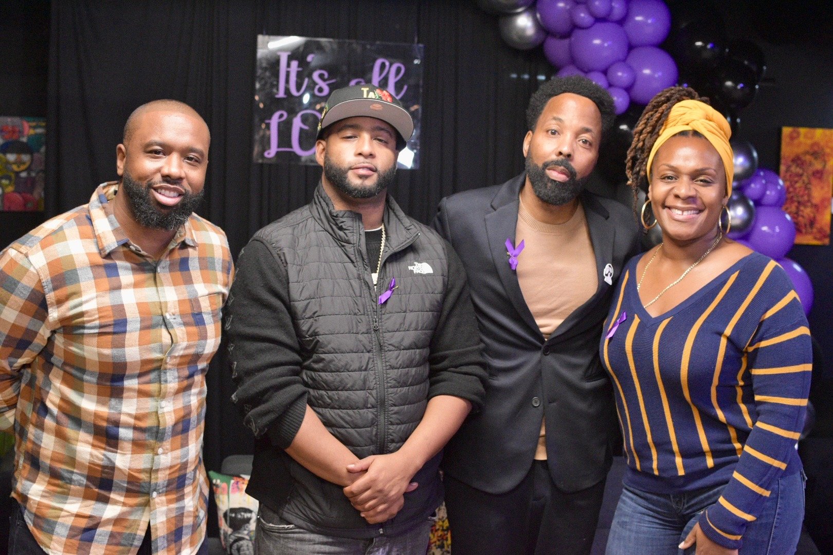 Four people smiling, standing in front of a background with purple balloons and a sign reading "It's all Love."