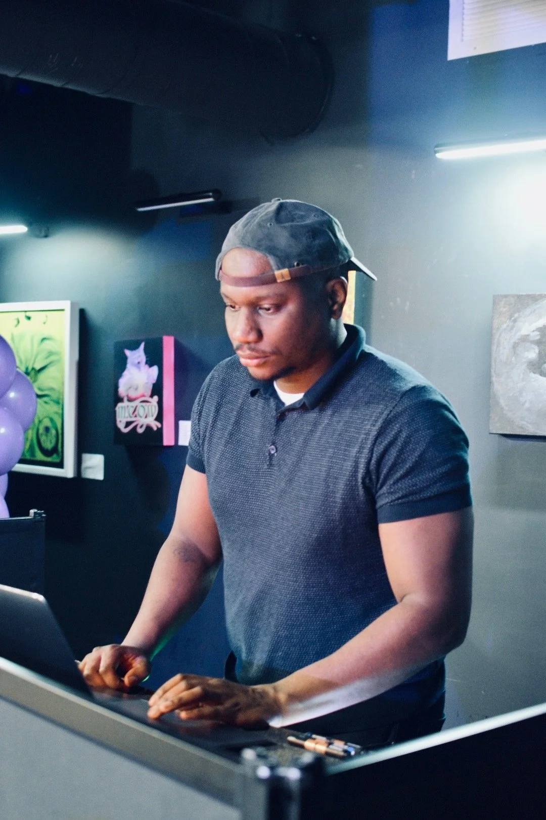 Man in a gray shirt and backward cap using a laptop in an art gallery with paintings on the wall.
