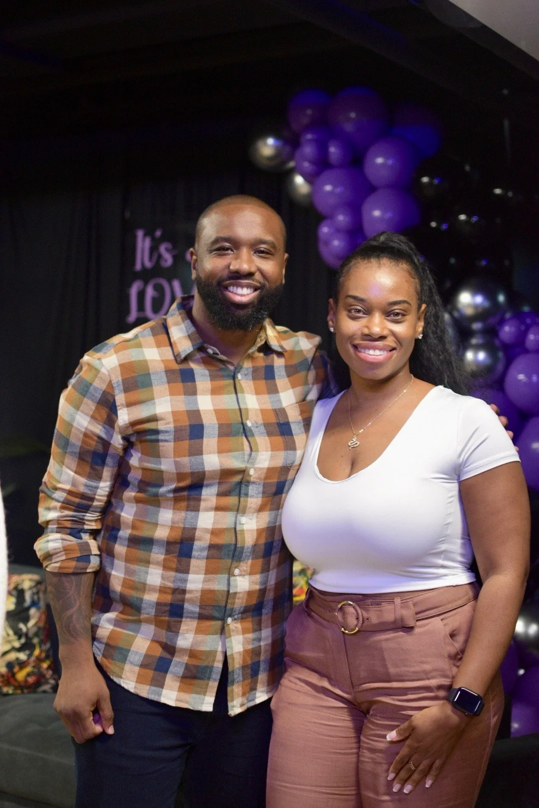 Man and woman smiling at a celebration with purple balloon decorations.
