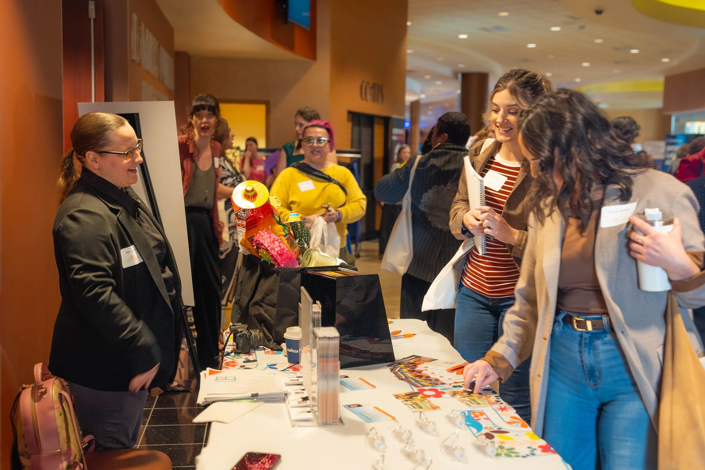 People engaging at a promotional or informational booth in a large indoor venue, with group conversations and displayed promotional materials on the table.