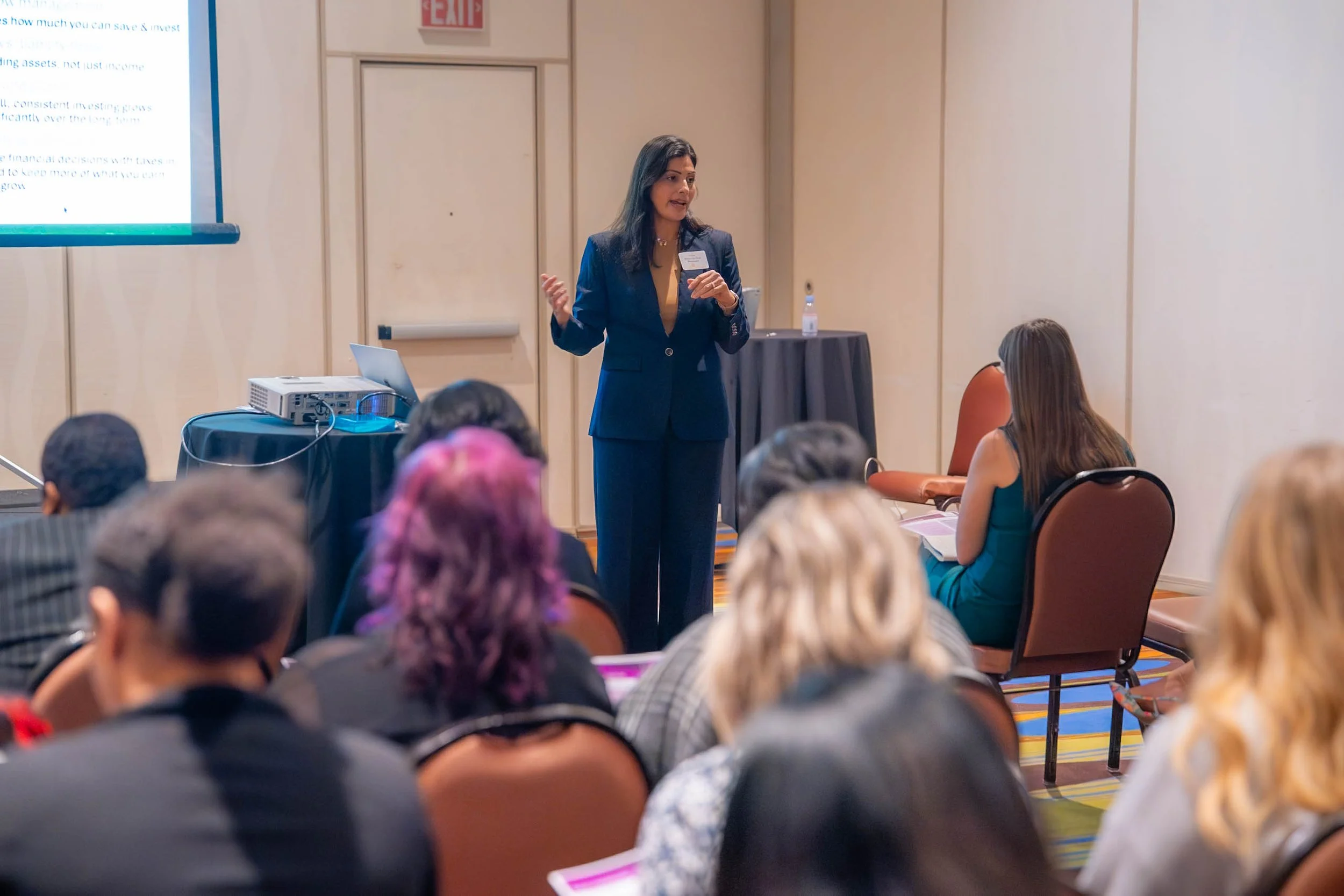 A woman in a dark blue blazer giving a presentation in a conference room with attendees seated and listening.