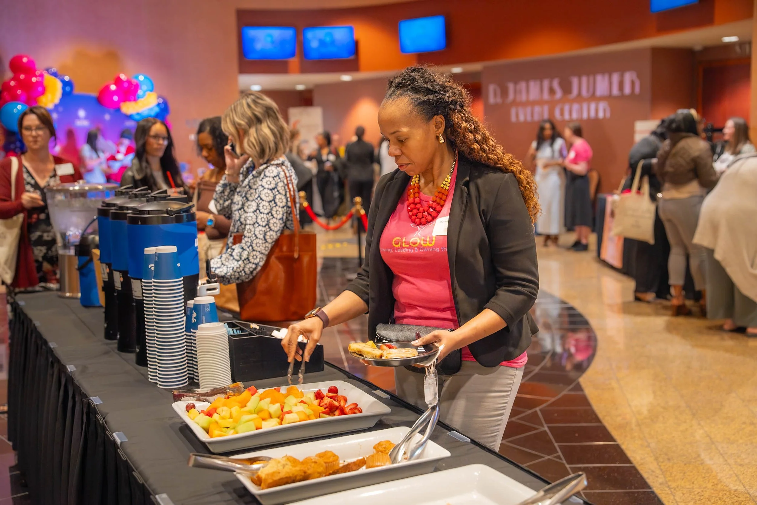 Woman selecting food at a buffet table with fruit, pastries, and hot food options; background features event signage and balloons.