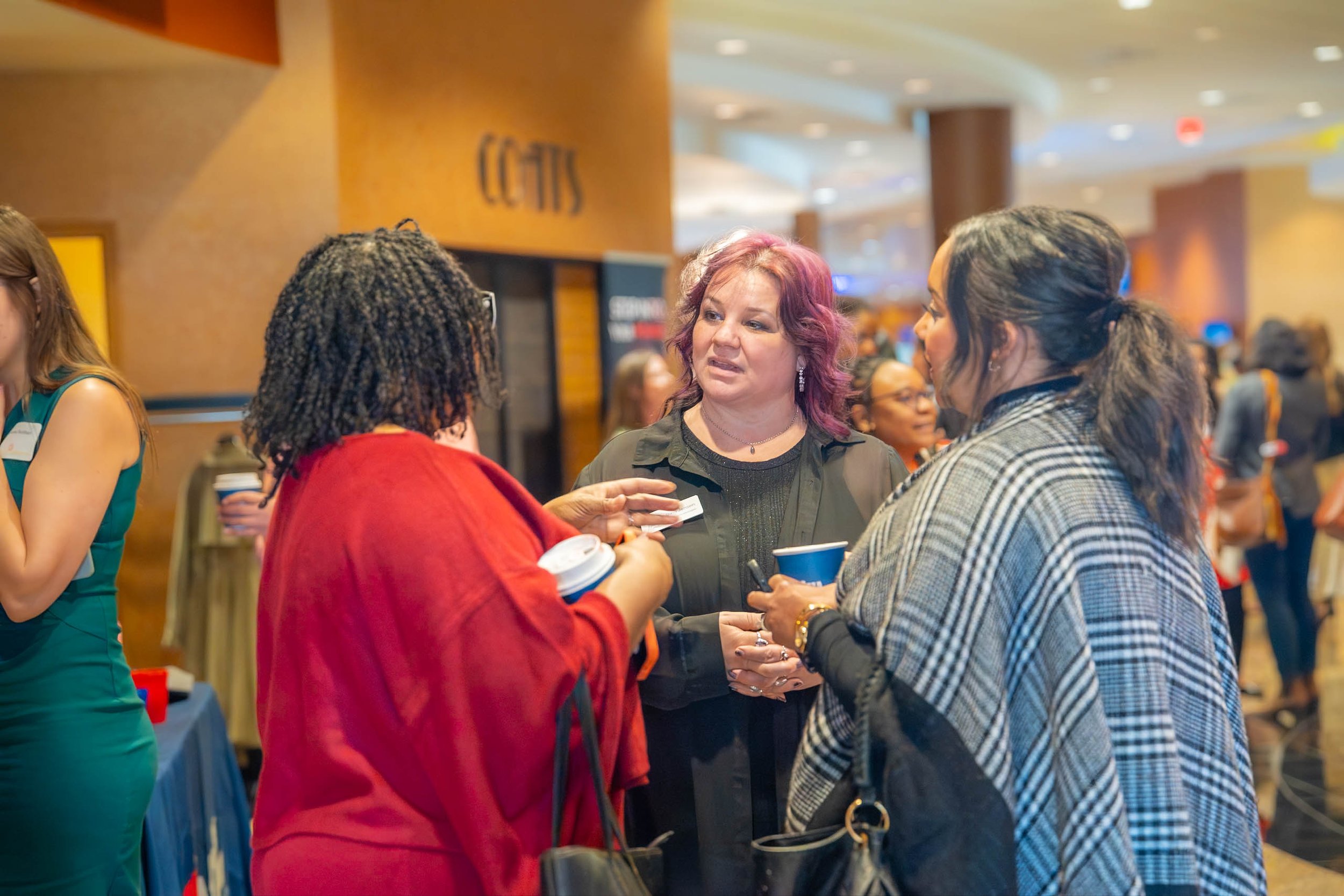 A group of women engaging in conversation at an indoor event. Four women are visible in the foreground, with one holding a coffee cup, and others listening and talking. The background shows a crowded hallway with informational signs.