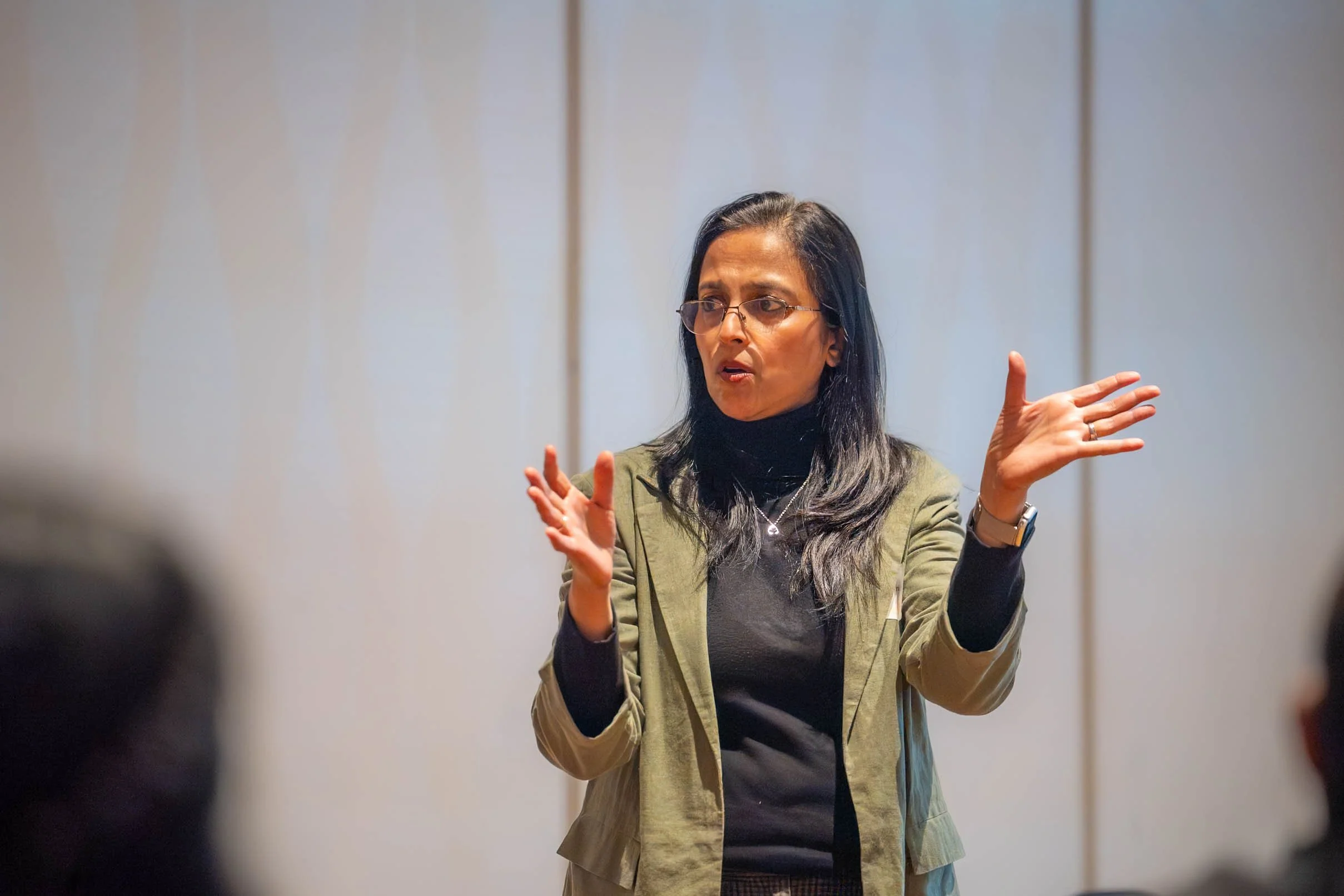 Woman with long dark hair, glasses, wearing a green jacket and black turtleneck, gesturing with her hands and speaking, on a plain wall background.