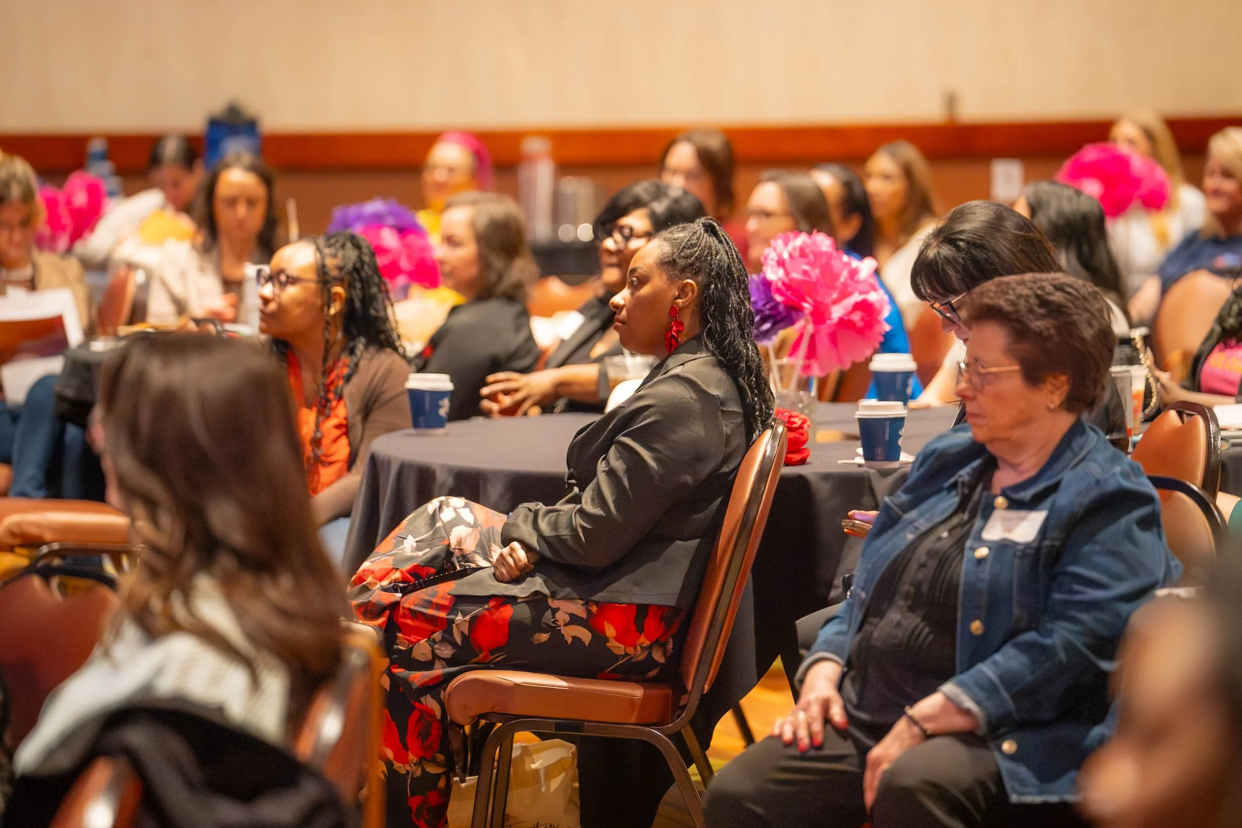 Group of women attending a conference or seminar, sitting at tables with pink and purple flower decorations, some wearing glasses, and holding cups.