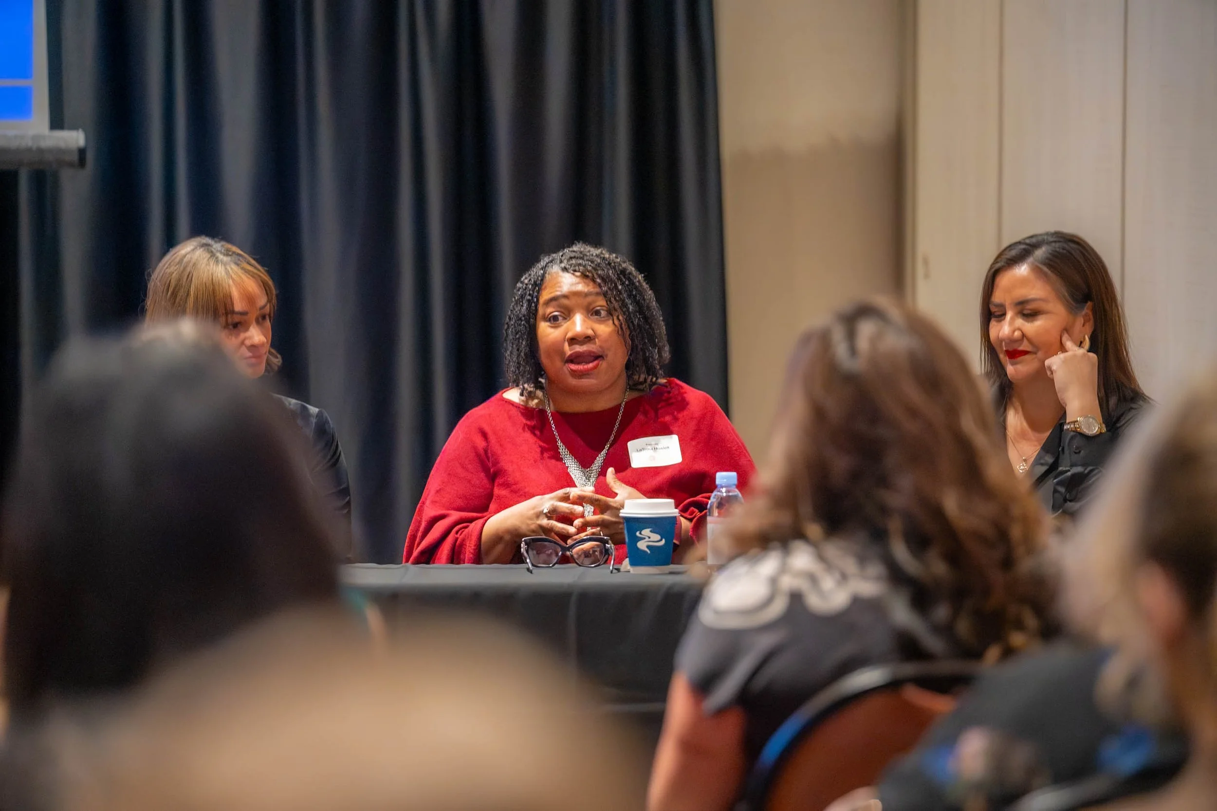 A group of women attending a conference or panel discussion, seated at a table, with one woman speaking and others listening, in an indoor setting with dark curtains in the background.