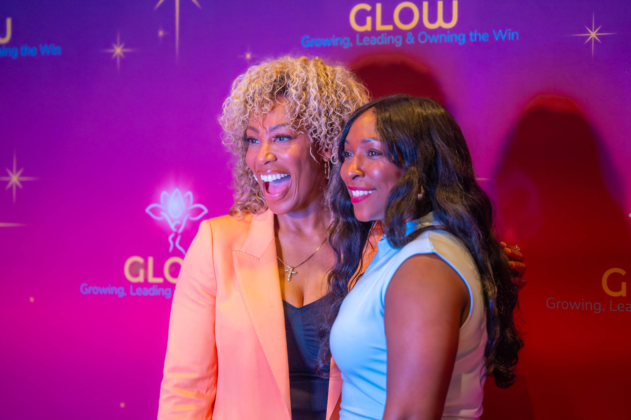 Two women standing together smiling in front of a pink and purple backdrop with the words 'Glow' and 'Growing, Leading & Owning the Win' visible. The woman on the left has curly blonde hair and is wearing a peach-colored blazer. The woman on the righ