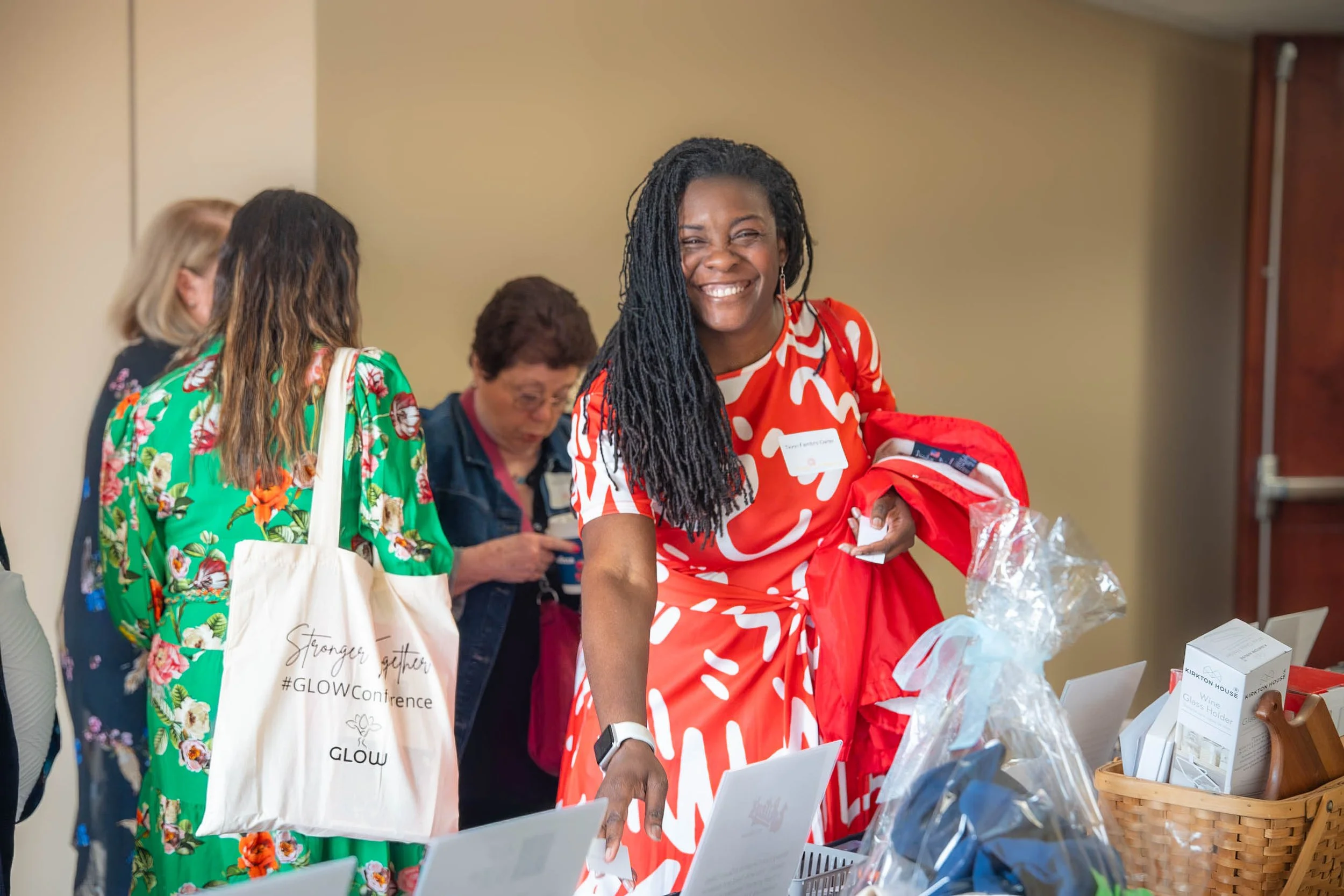 A smiling Black woman with long dreadlocks wearing a red and white patterned dress, holding a red bag, at a table with various items for sale or display, in an indoor setting.