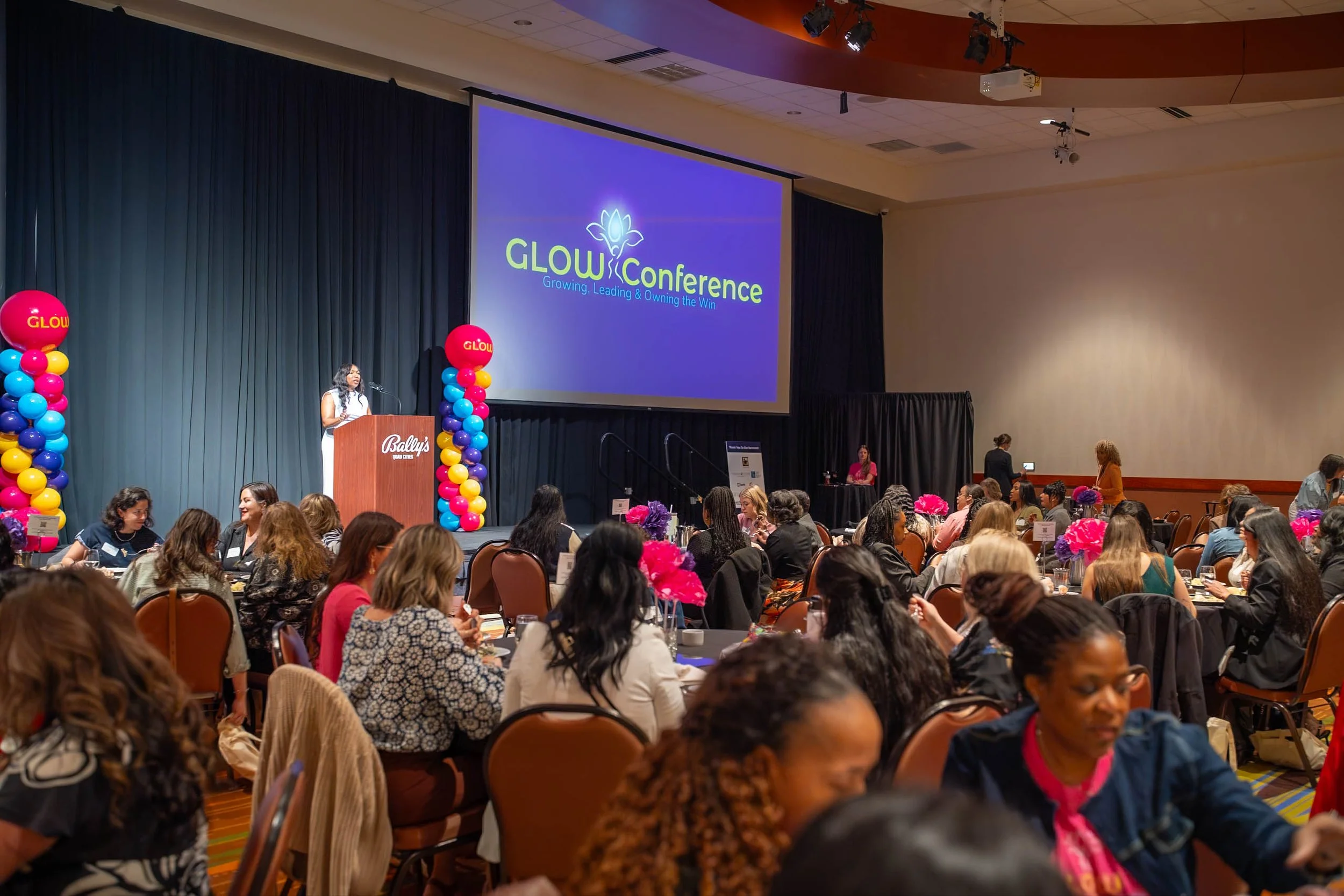 A conference room filled with people seated at tables decorated with pink and purple flowers. There is a stage with a speaker at the podium and a large screen displaying the logo 'GLOW Conference' with the slogan 'Growing, Leading & Owning the Win.' 