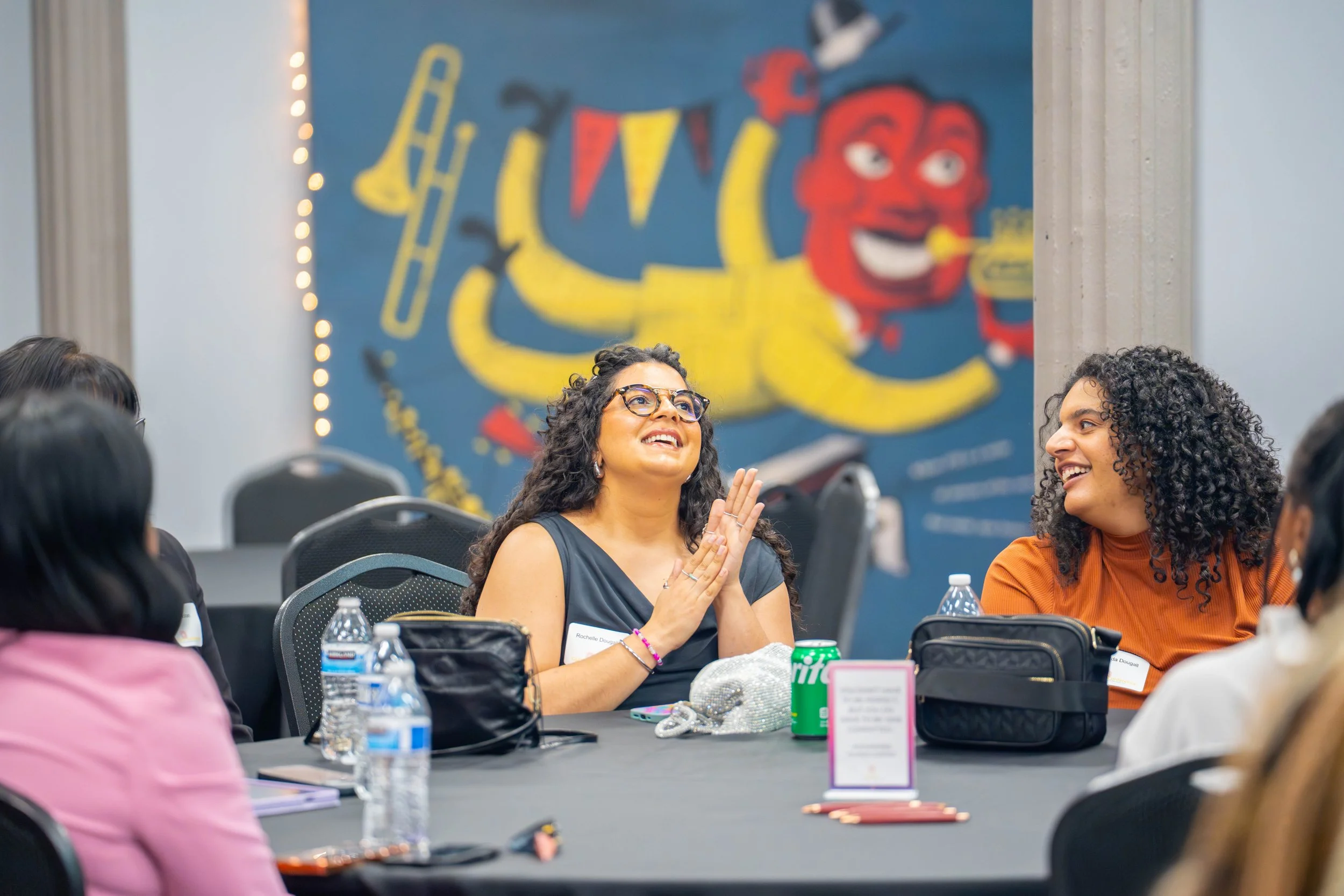 Two women seated at a conference table, smiling and clapping, with water bottles and personal items in front of them, in a room decorated with a colorful mural of a friendly red character and yellow objects on the wall behind them.