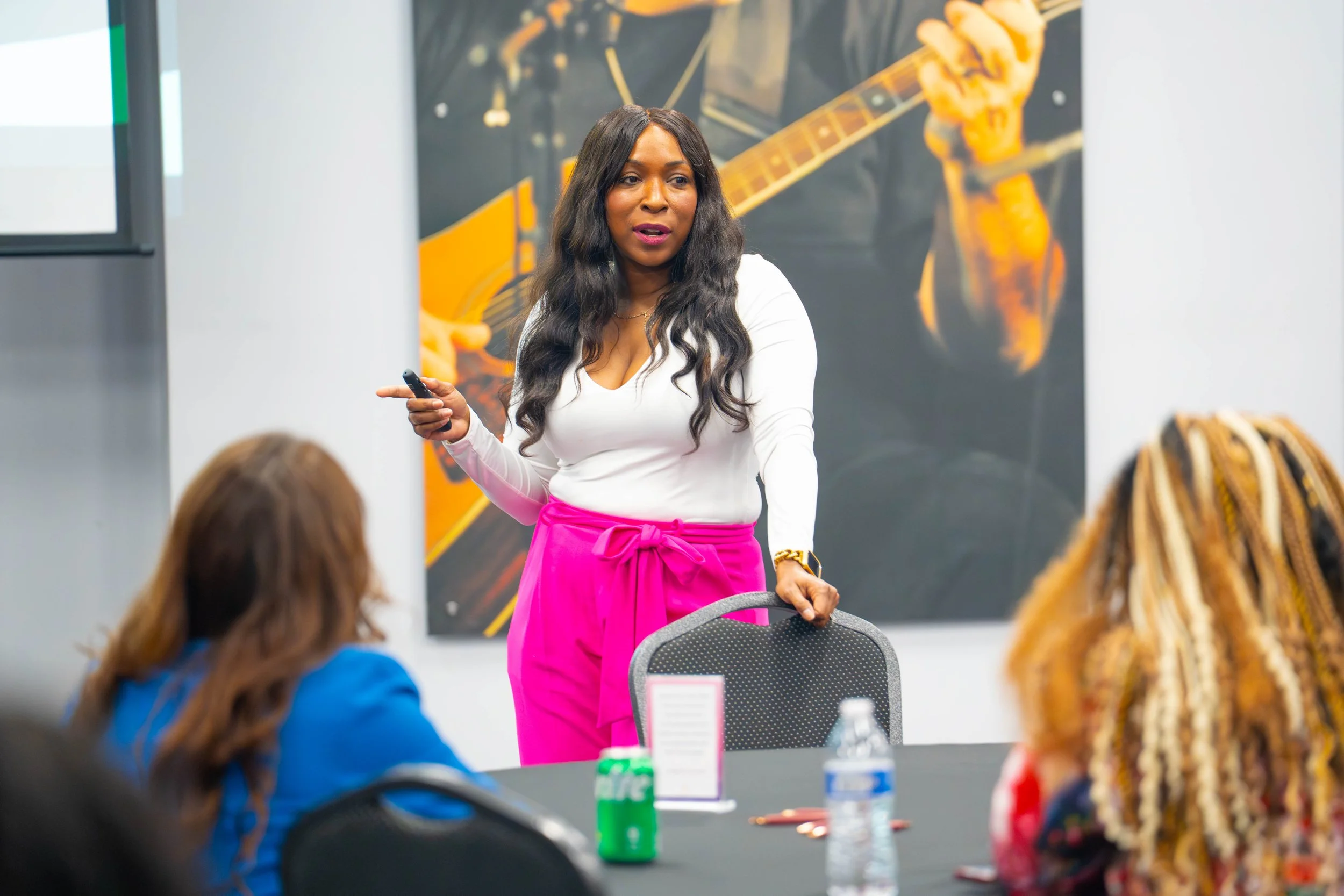 A woman with long black wavy hair, wearing a white top and bright pink high-waisted pants, is standing in front of a group, holding a remote or laser pointer, speaking to an audience in a conference room with a large poster of a person playing a guit