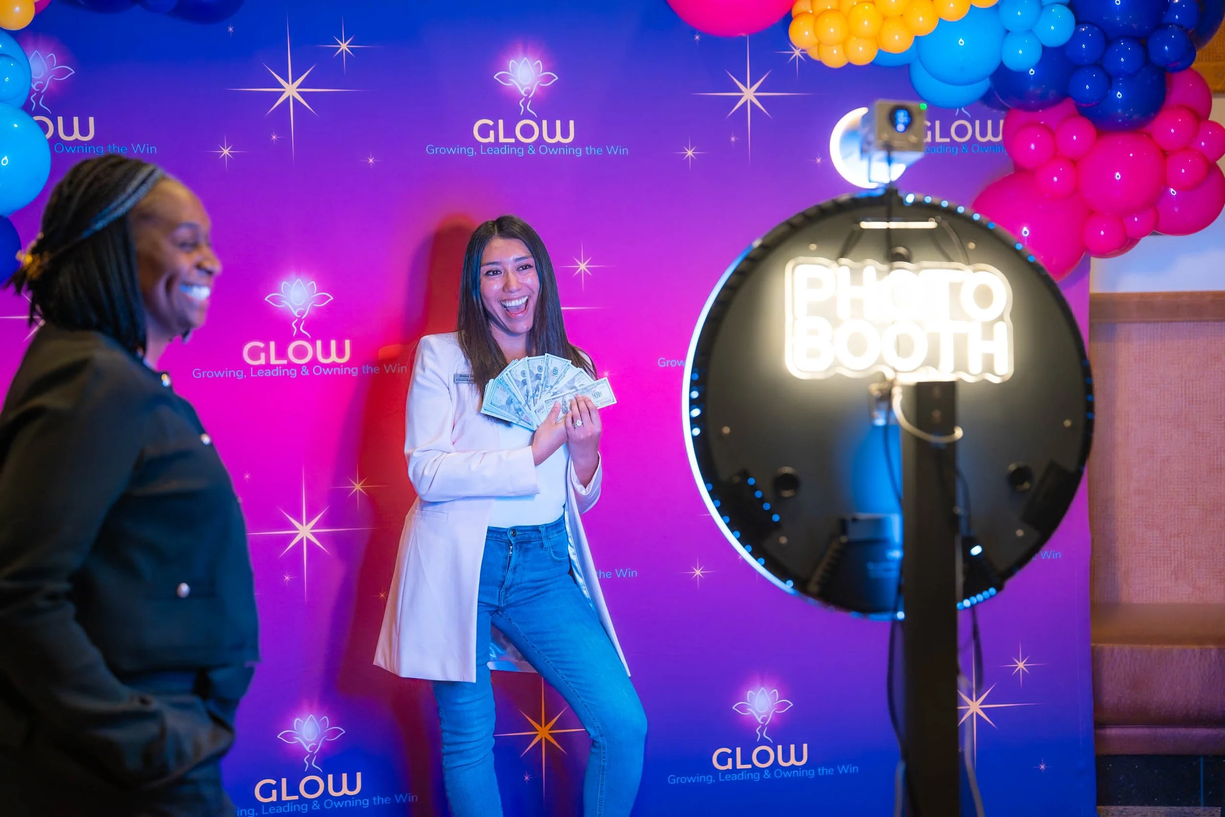 Two women at a photo booth with a purple background decorated with balloons in pink, blue, and yellow, and the word 'GLOW' with the tagline 'Growing, Leading & Owning the Win.' One woman, smiling, holds a fan of cash, while the other woman is smiling