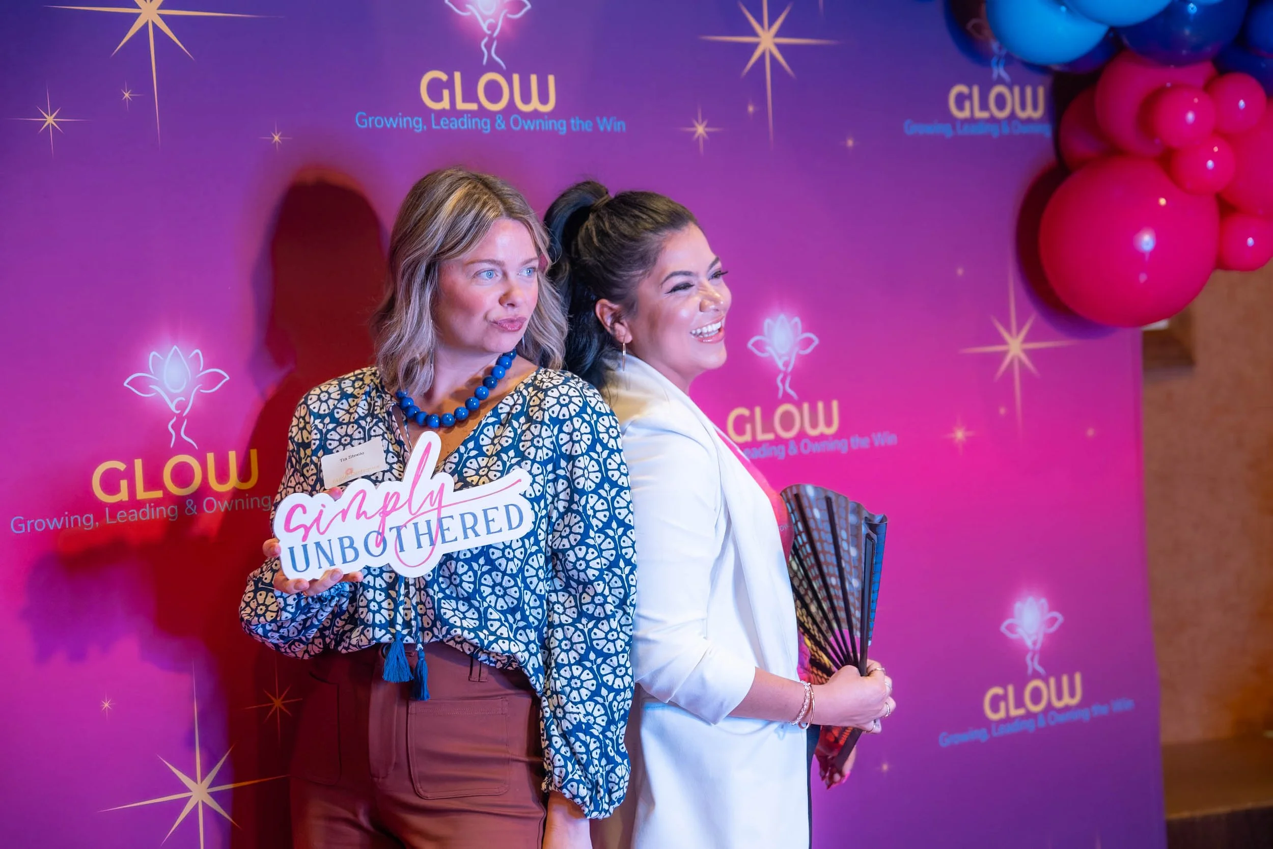 Two women standing in front of a pink and purple backdrop with balloons. One woman is holding a sign that says "Simply Unbothered." The backdrop features the word "GLOW" and the slogan "Growing, Leading & Owning the Win." Both women are smiling and l