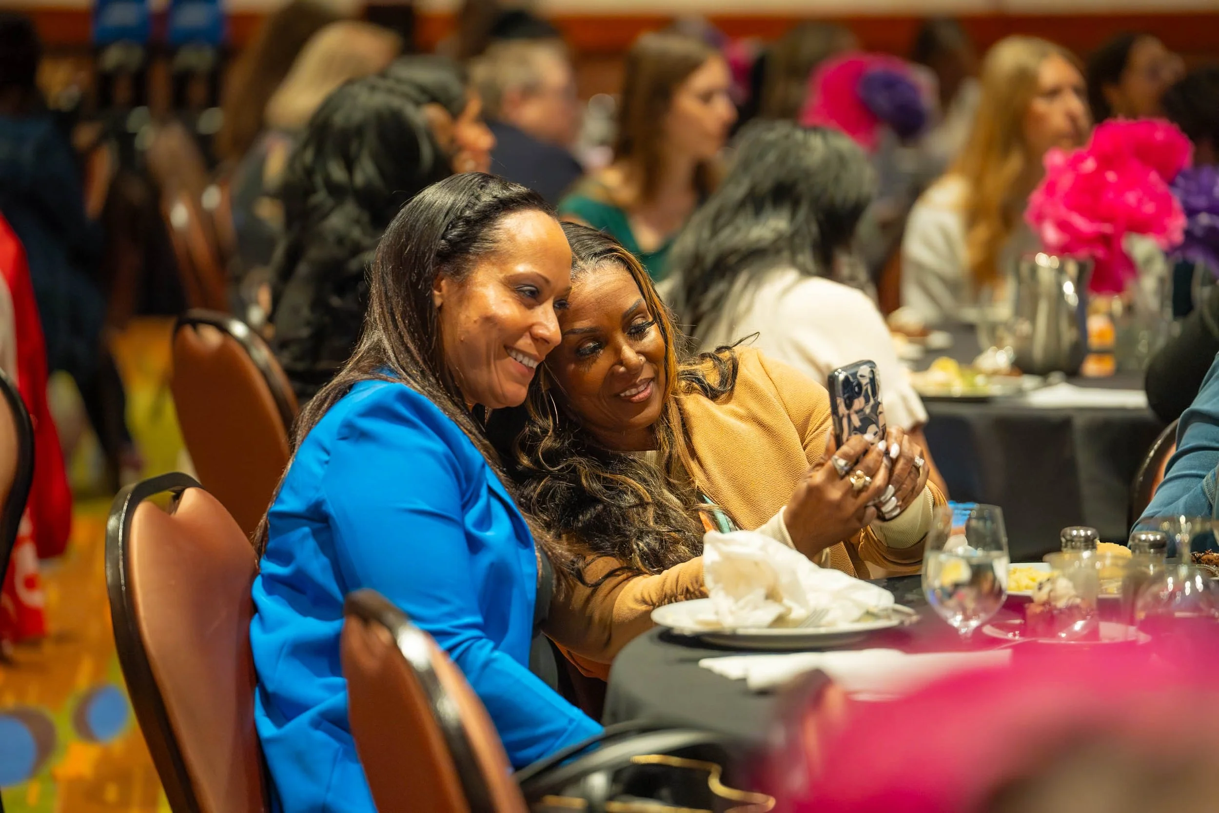 Two women sitting closely at a table, looking at a phone together, smiling, during a social or formal event with multiple people in the background.