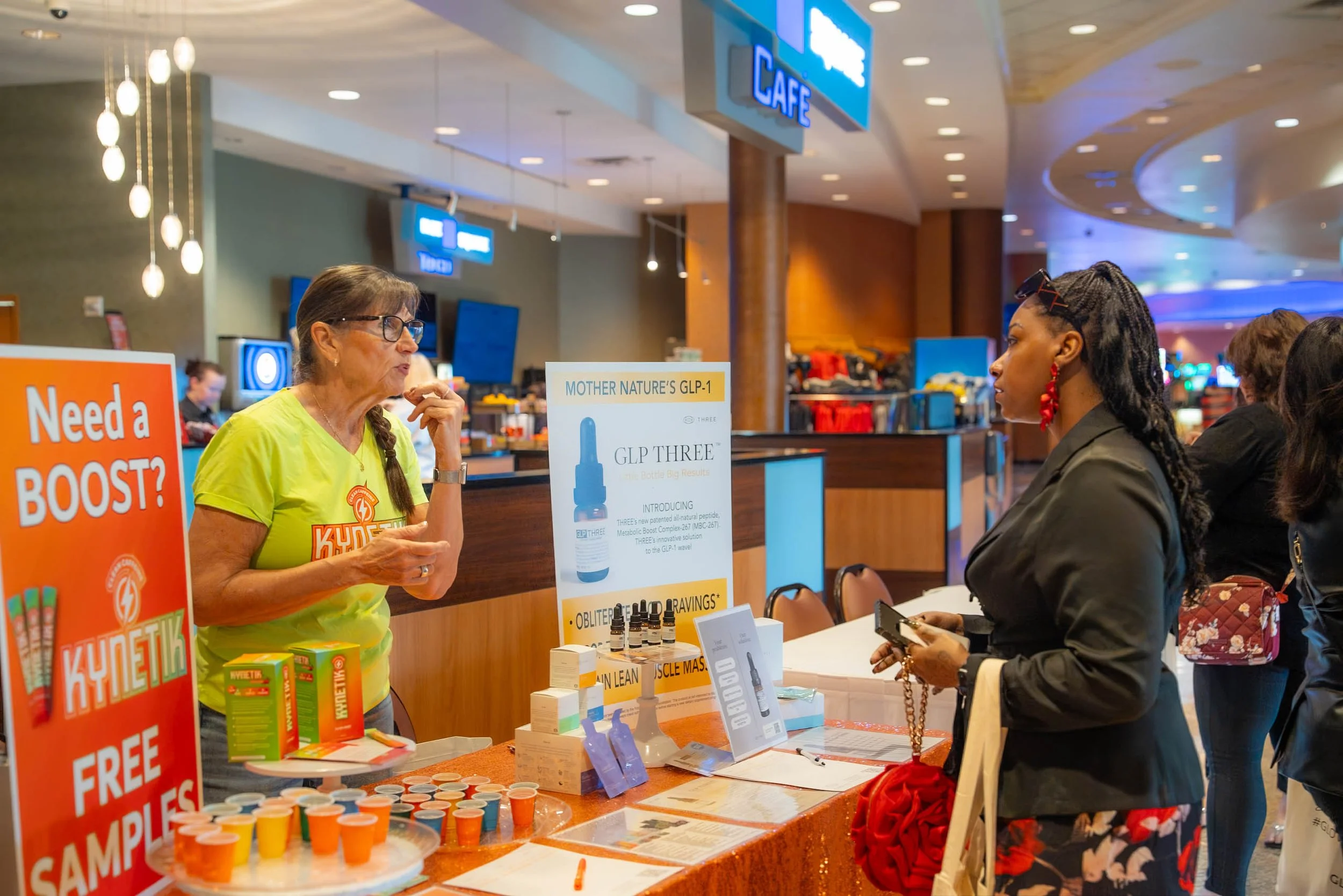 A woman in a lime green shirt is talking to a woman in a black jacket at a booth in a shopping mall or event space. The booth has various health or beauty products on display, including small bottles, samples, and promotional signs. Behind the woman 