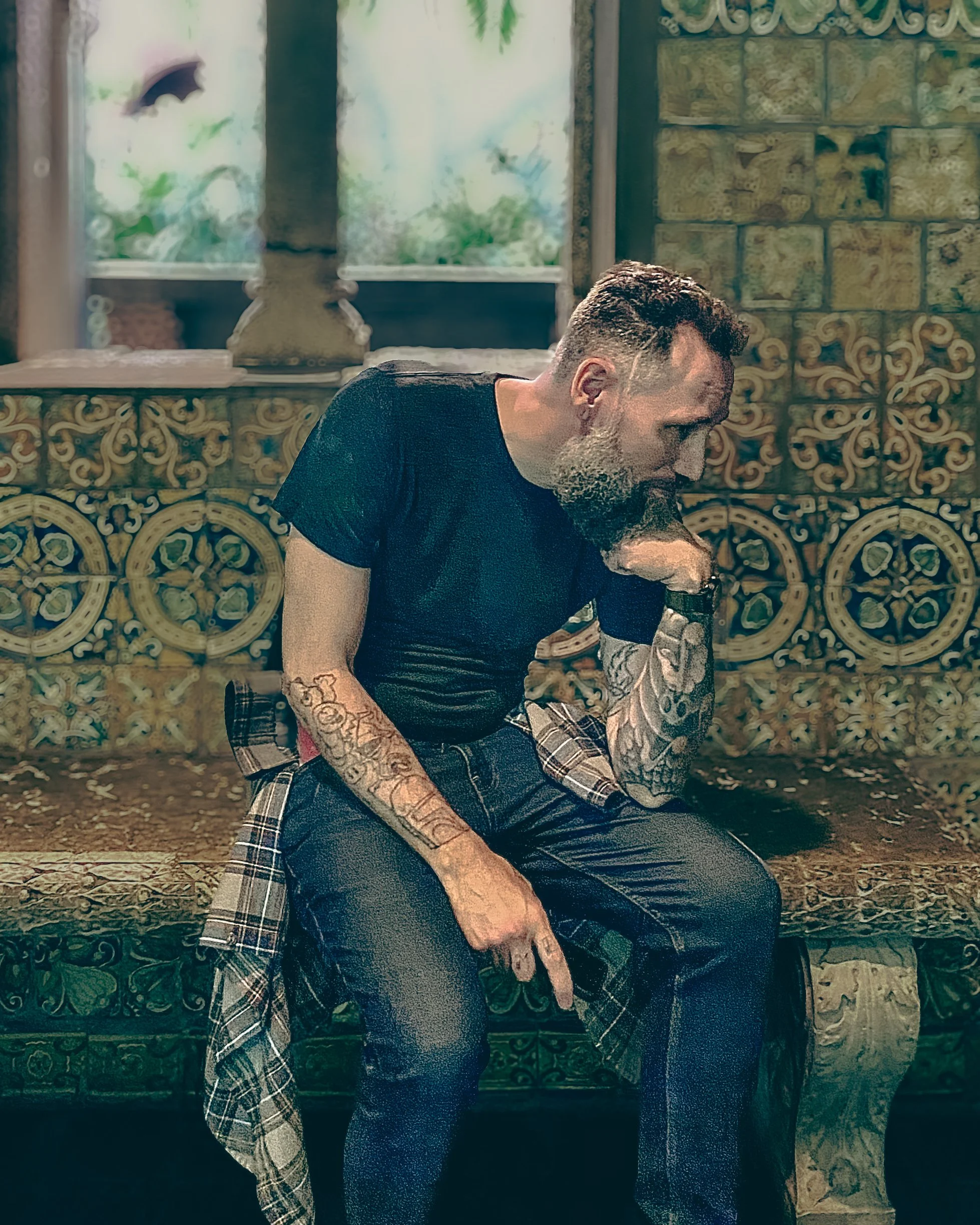 A man with tattoos and a beard sitting on a decorative bench, resting his chin on his hand in a contemplative pose inside a room with ornate wall decor and a window with a view of trees.