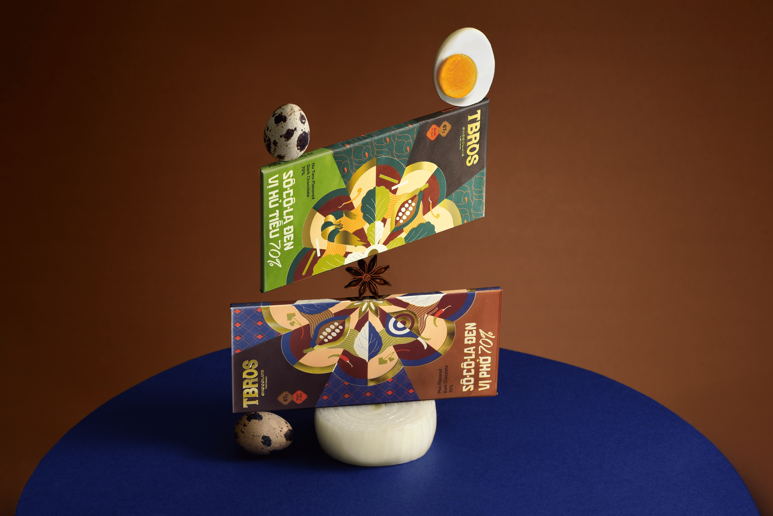 Artistic still life of two chocolate bar boxes, a white onion, a quail egg, and a half-cut boiled egg, balanced on a black star-shaped object, against a brown background with a blue circular surface.