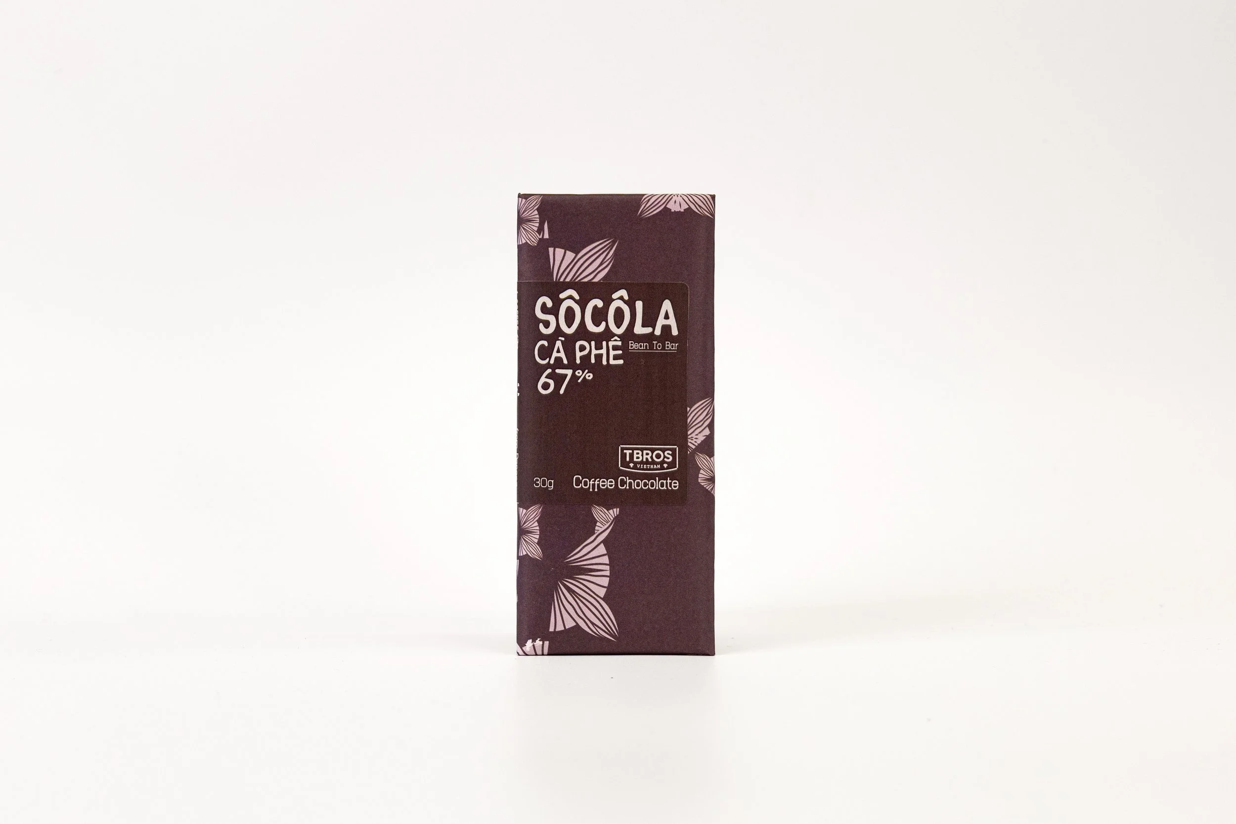 A dark brown chocolate bar with floral designs and text indicating it is a coffee-flavored bean to bar chocolate, 67% cacao content, weighing 30 grams, from the brand T BROS.