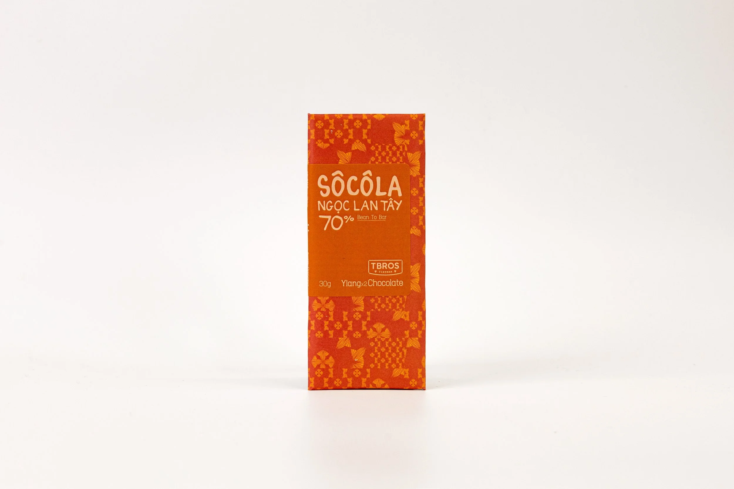 A rectangular chocolate bar package with predominantly orange and red colors, decorated with floral patterns, with text indicating "Sôcôla Ngọc Lan Tây 70%" and other details in Vietnamese.