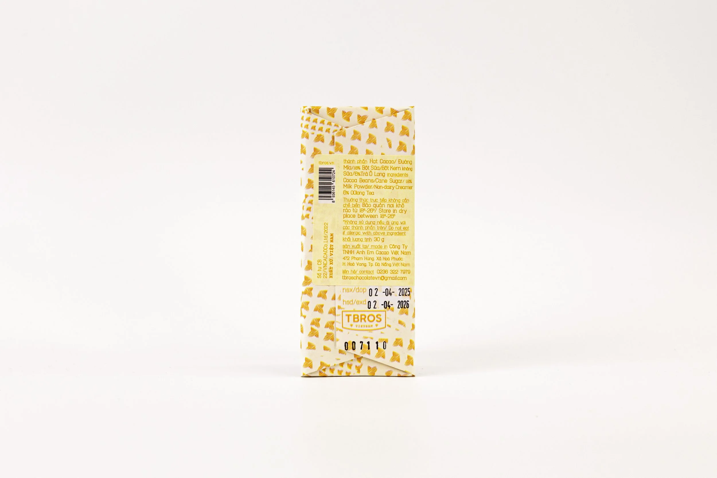A small, rectangular chocolate bar package with a yellow and white patterned design, featuring a yellow label with product information and expiration dates.