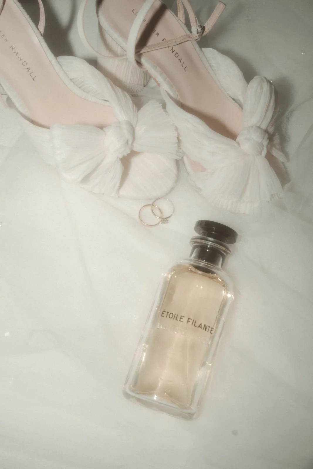 Ein Paar weißer High Heels mit Schleifen, ein Parfümflasche namens 'Étoile Filante', zwei goldene Ringe und ein Paar goldene Eheringe auf weißem Stoff