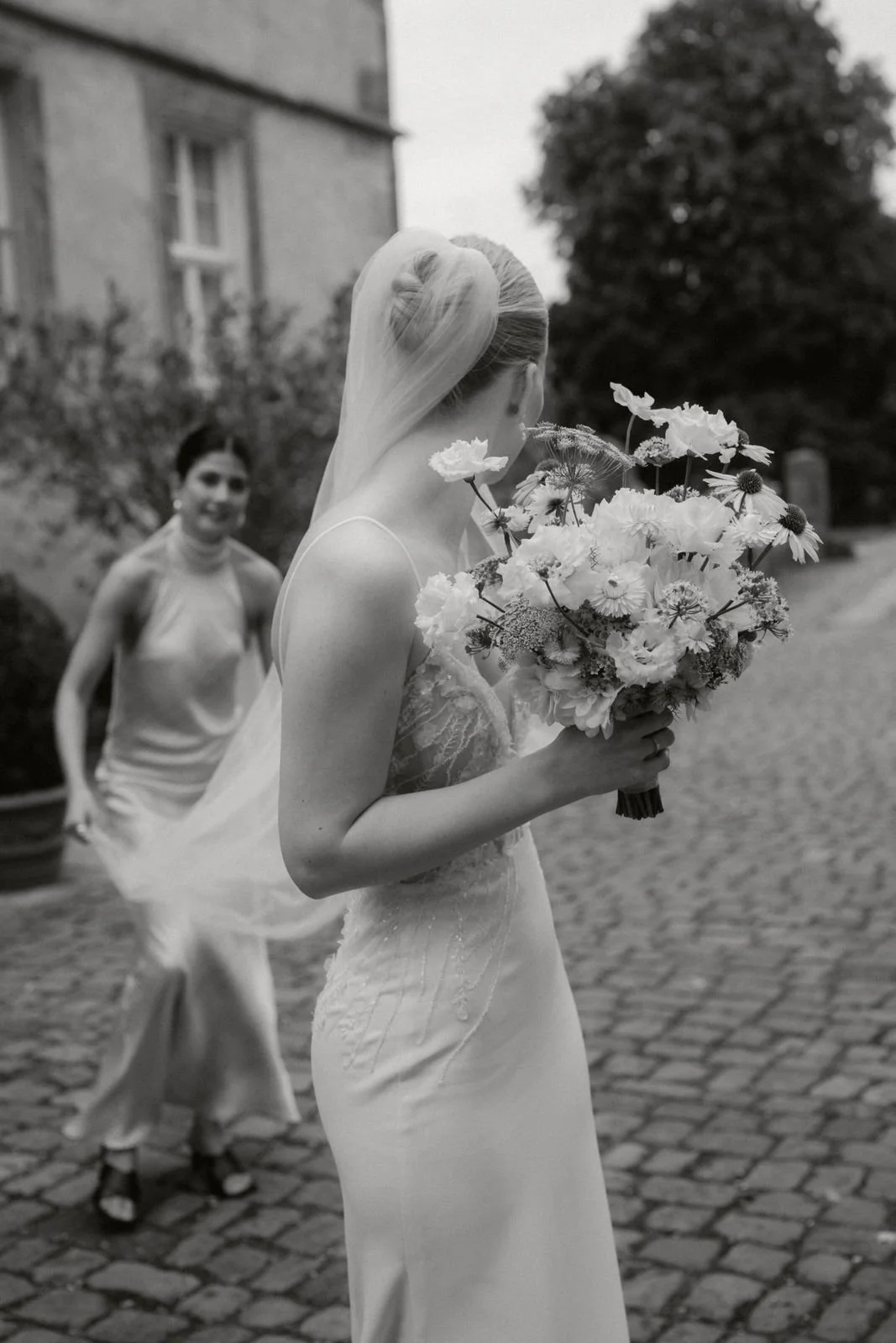 Braut mit Hochzeitsstrauß in der Hand, im Vordergrund, und eine Frau im Abendkleid im Hintergrund, auf einer gepflasterten Straße, im Freien.